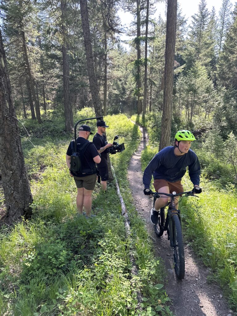 A mountain biker goes past a two-person film crew. They are in the deep woods on a sunny day.