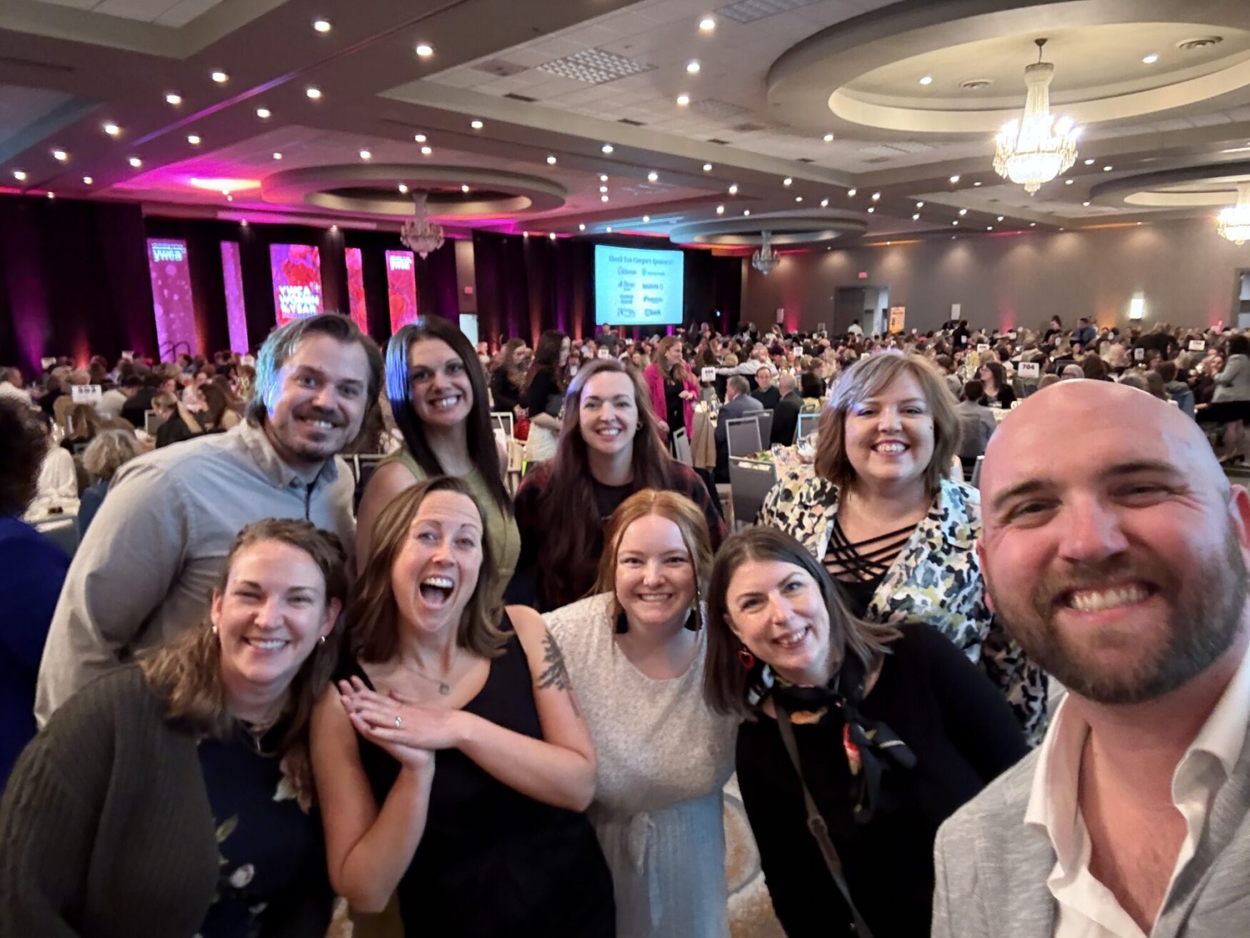 A cheerful group of ten adults poses for a selfie at a lively banquet event in a grand ballroom. The background shows a large crowd seated at round tables, with chandeliers above and pink stage lights illuminating banners that read “YWCA Women of the Year.” Everyone in the group is smiling, with some striking playful poses, dressed in business casual and semi-formal attire.