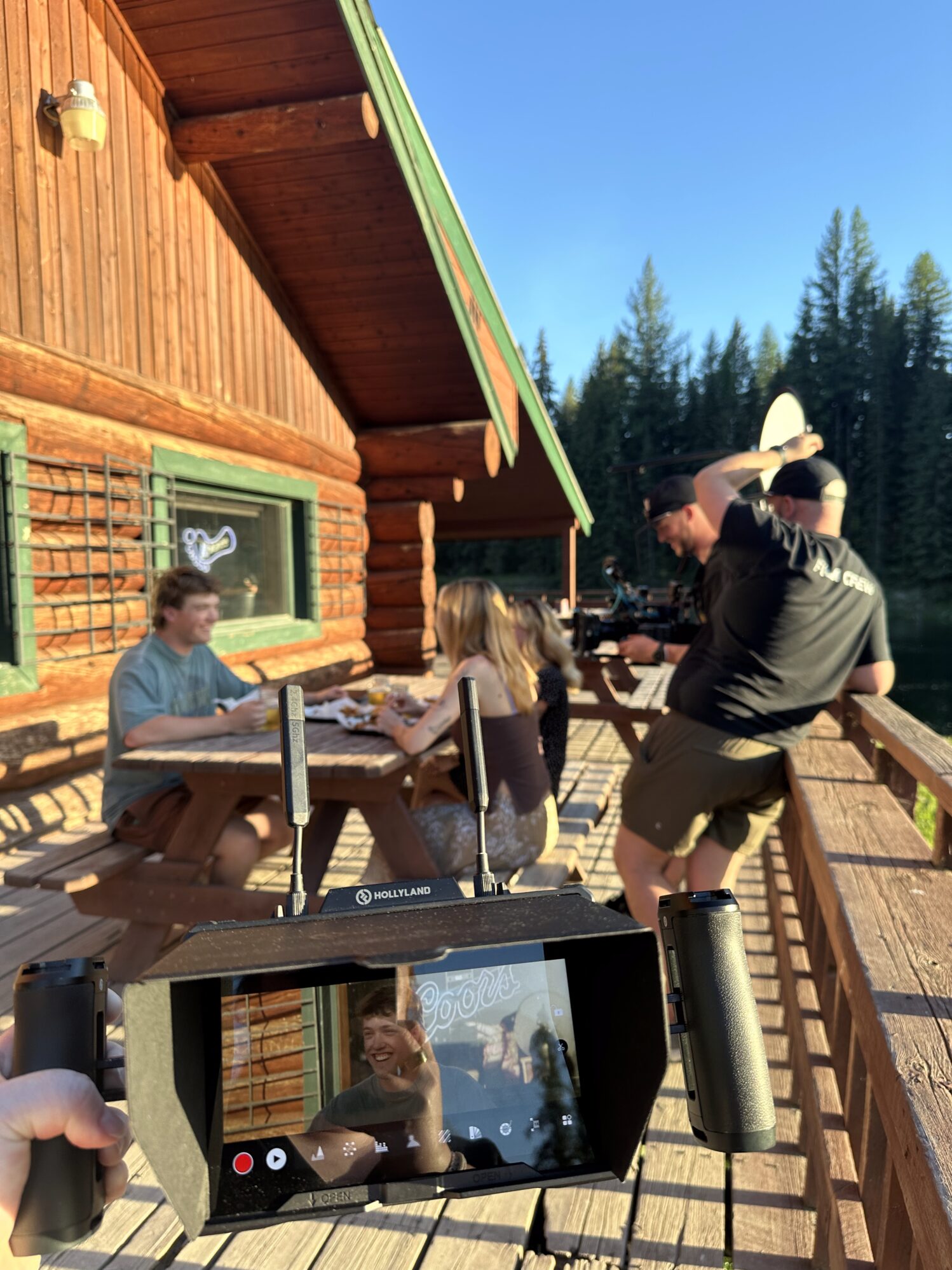 Three people sit at a picnic table with a two-person film crew filming them. Another person is holding a director's monitor in the foreground, watching the filming happen. It is sunny and on an outdoor patio.