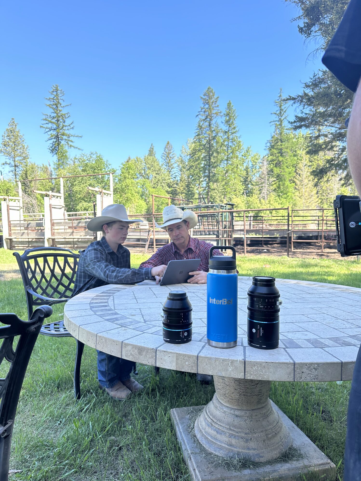 Two men in cowboy hats sit a stone table watching something on a tablet. There is a camera man pointing a camera at them. On the table is a water bottle that says "InterBel" and two additional camera lenses.