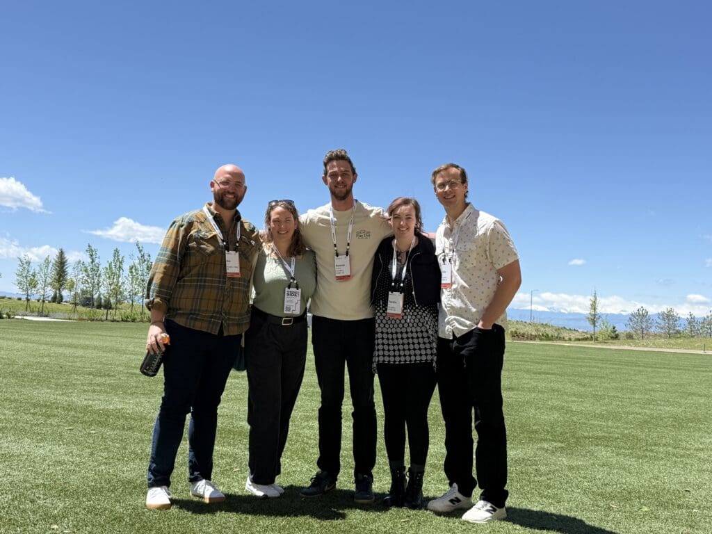 Five people stand closely together, smiling, on a sunny day in a wide open grassy field with trees and mountains in the background. They are wearing name tags and conference badges, suggesting they are attending an event. The sky is clear and blue. Everyone is dressed casually, and the atmosphere appears friendly and relaxed.