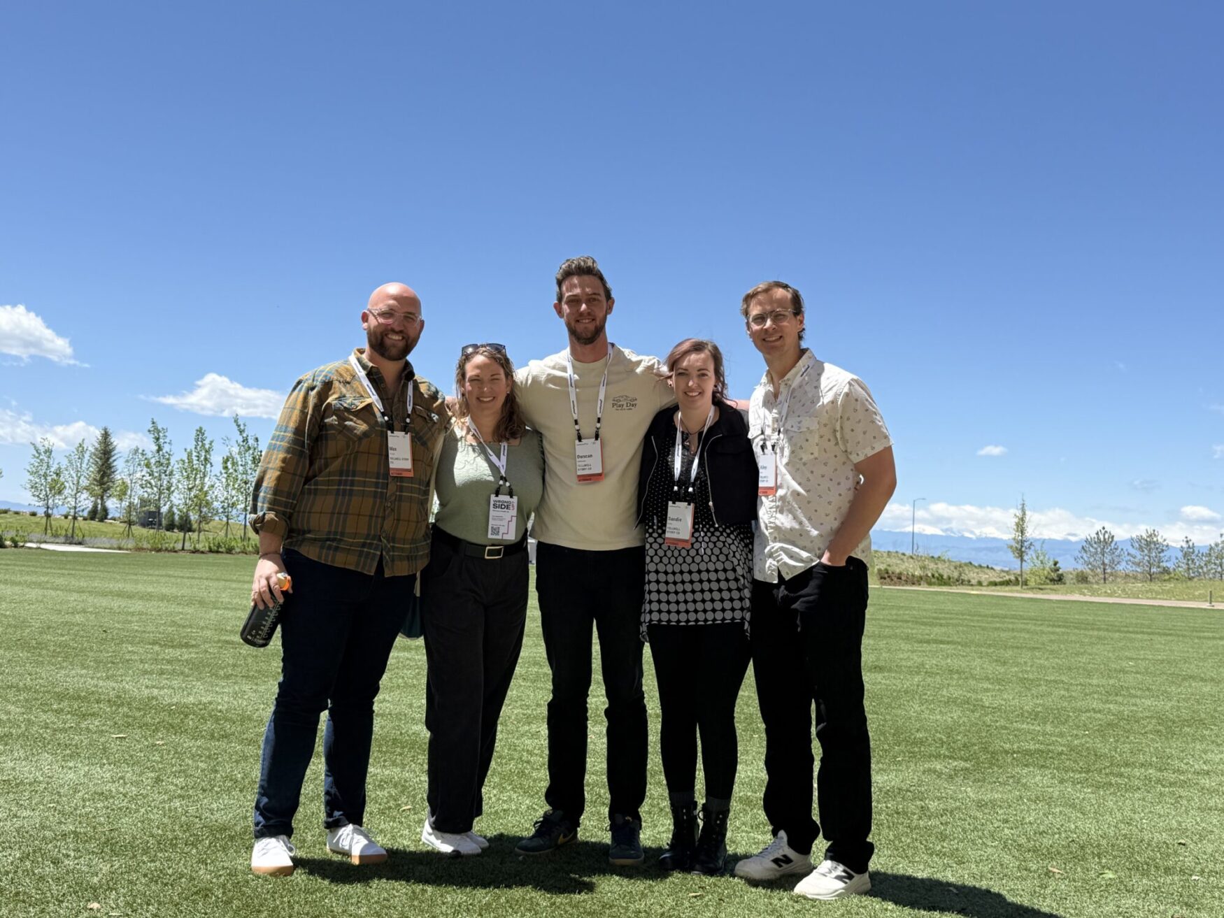 Five people stand closely together, smiling, on a sunny day in a wide open grassy field with trees and mountains in the background. They are wearing name tags and conference badges, suggesting they are attending an event. The sky is clear and blue. Everyone is dressed casually, and the atmosphere appears friendly and relaxed.
