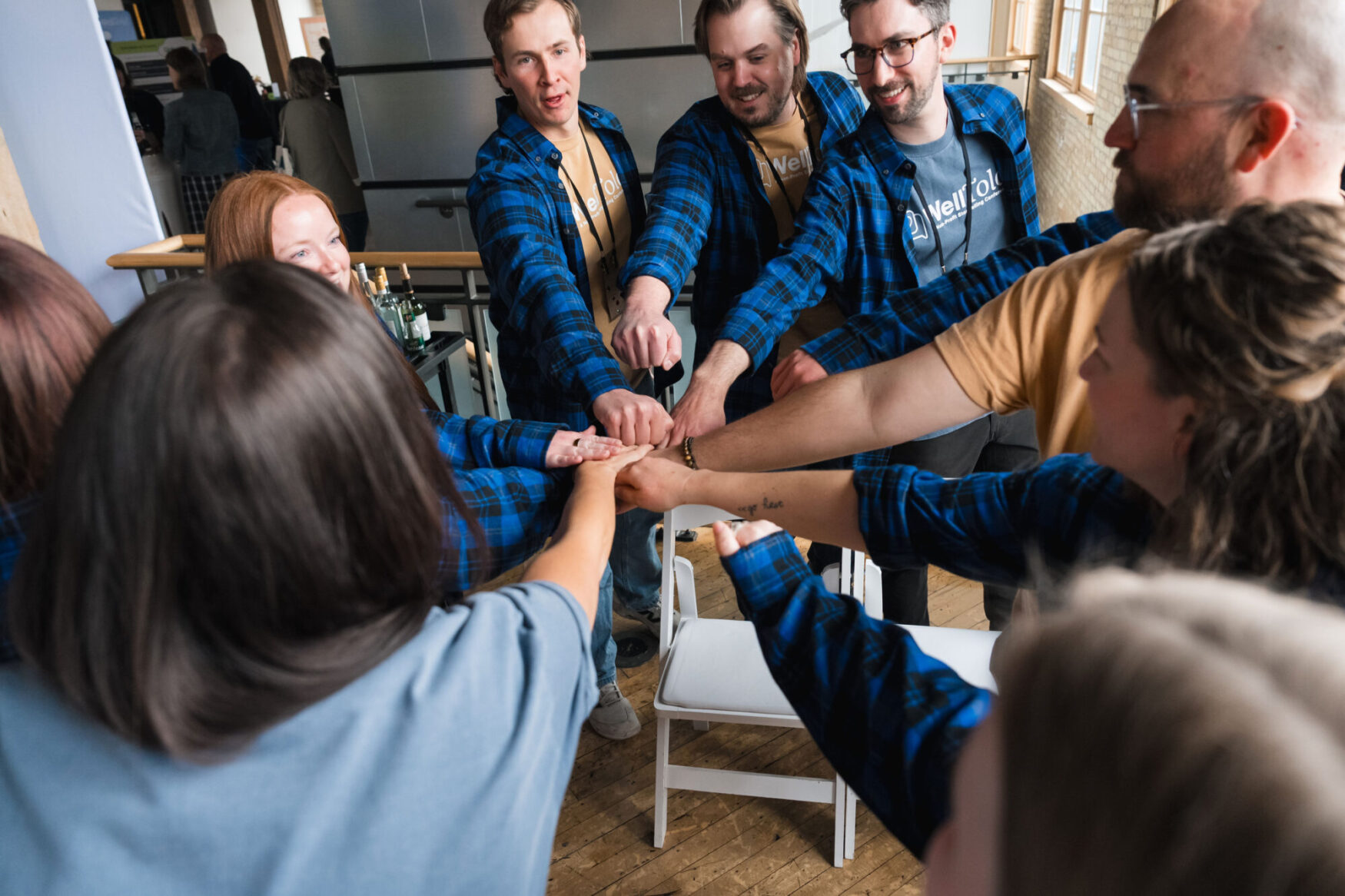 A group of men and women with their hands all in a circle, they look like they are going to cheer. They are all wearing blue and black plaid flannels.