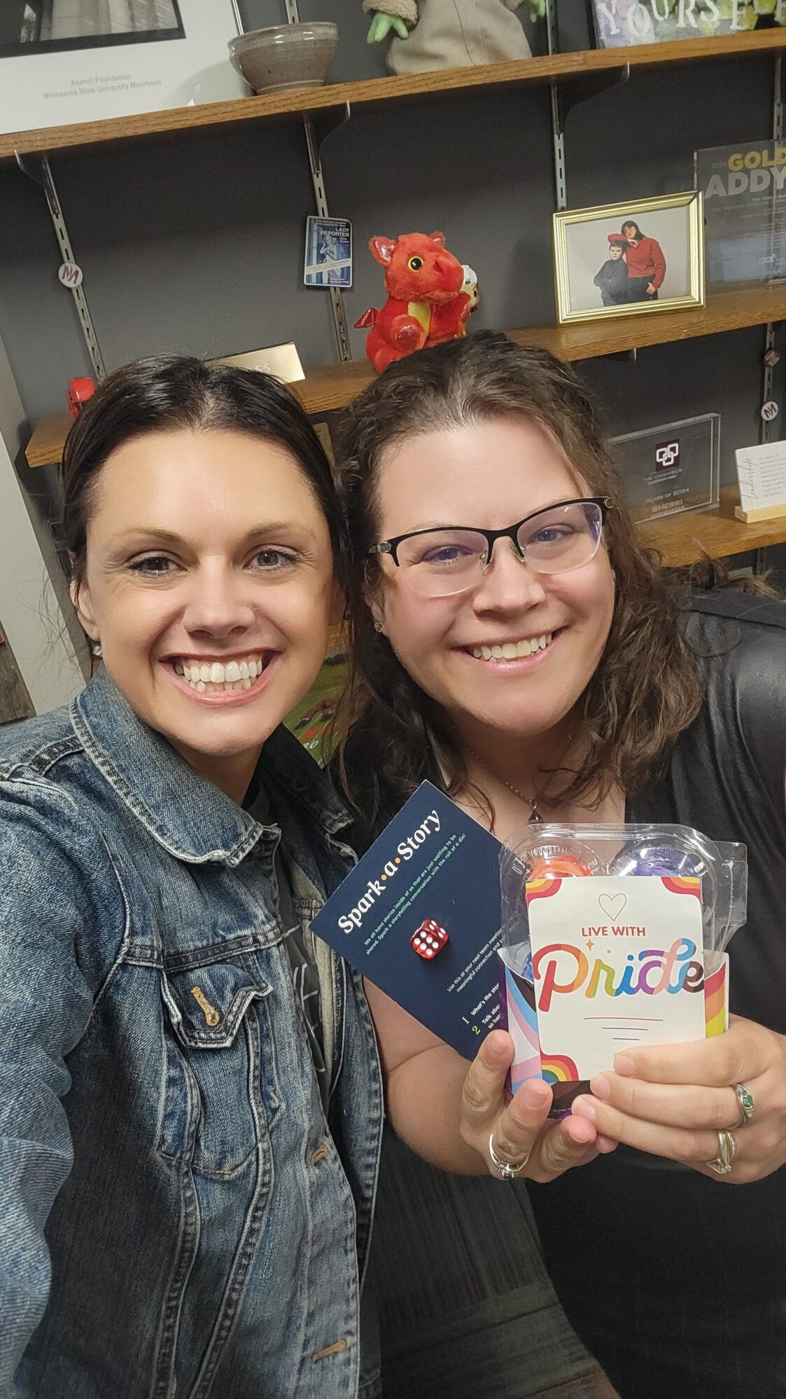 Two women smile for a selfie in an office space, holding a Pride-themed treat pack and a Spark-a-Story card with a red die, surrounded by shelves with personal items and awards.