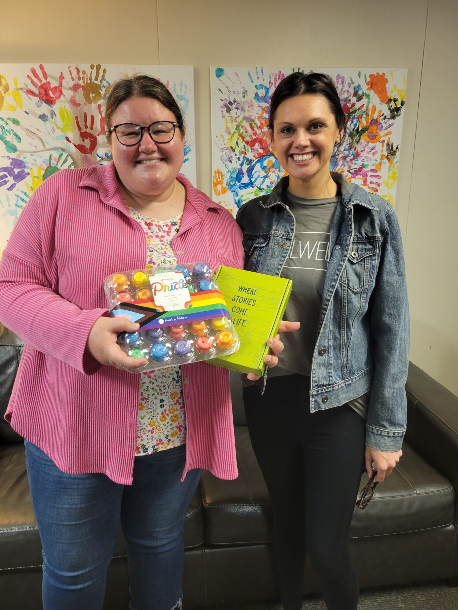 Two women smile in front of colorful handprint art, with one holding a set of vibrant paints and a bright green box labeled “Where Stories Come to Life.”