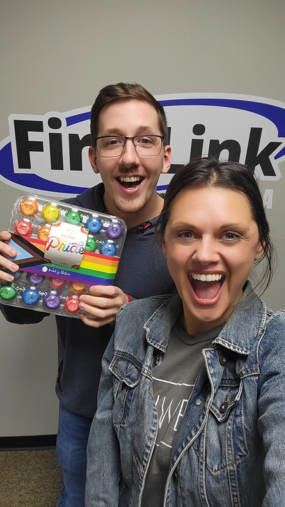 Two people smile excitedly in front of a FirstLink sign, holding a colorful tray of mini cupcakes decorated with rainbow Pride-themed packaging.