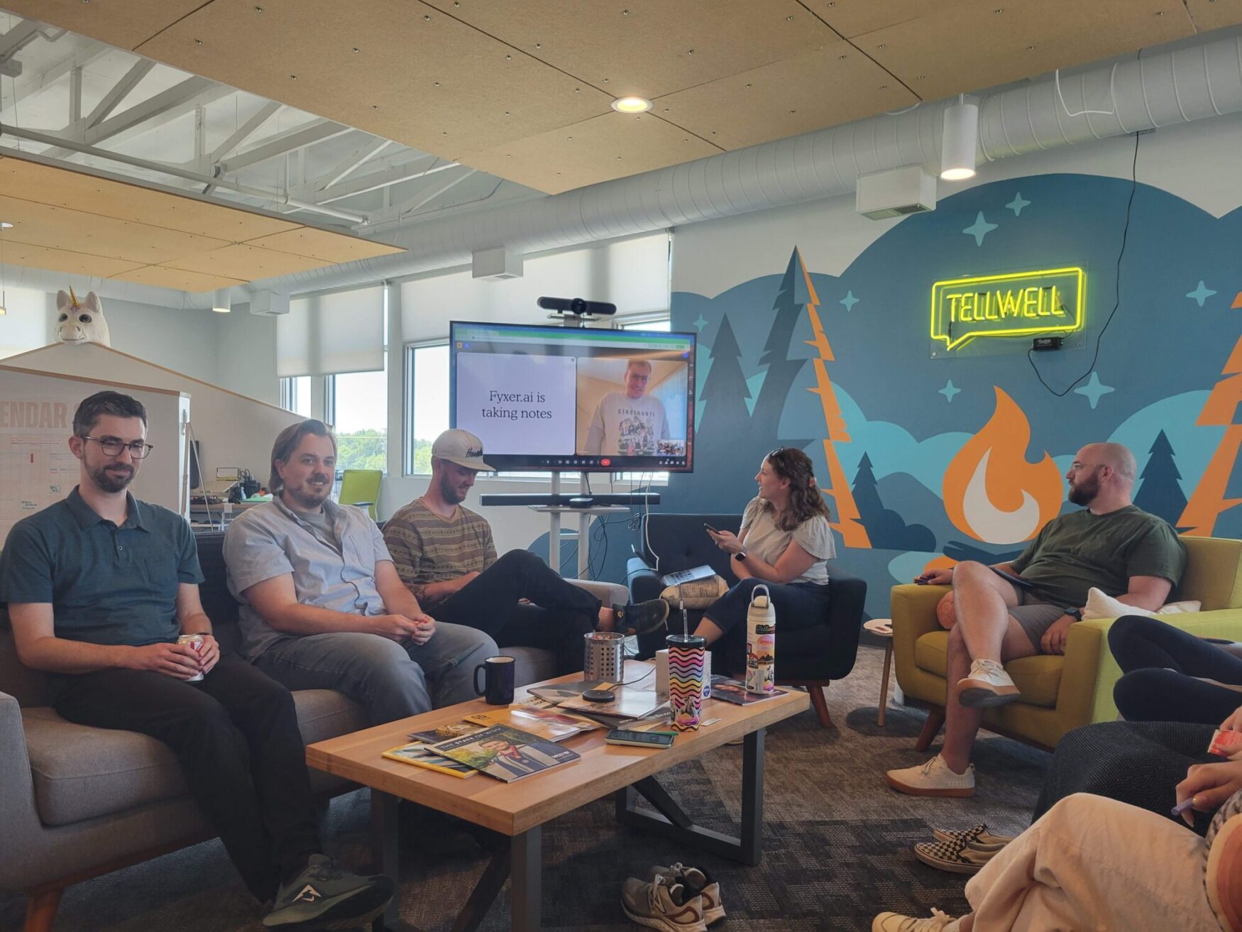 A group of people is seated in a modern office lounge, participating in a meeting. They are sitting on couches and armchairs arranged in a semicircle around a large screen. The screen displays a video call and shows a person speaking remotely. The room has colorful wall art featuring trees, mountains, and a campfire, with a neon sign that reads "TELLWELL" mounted on the wall. A coffee table in the center holds magazines, mugs, and water bottles. The atmosphere appears casual and collaborative.