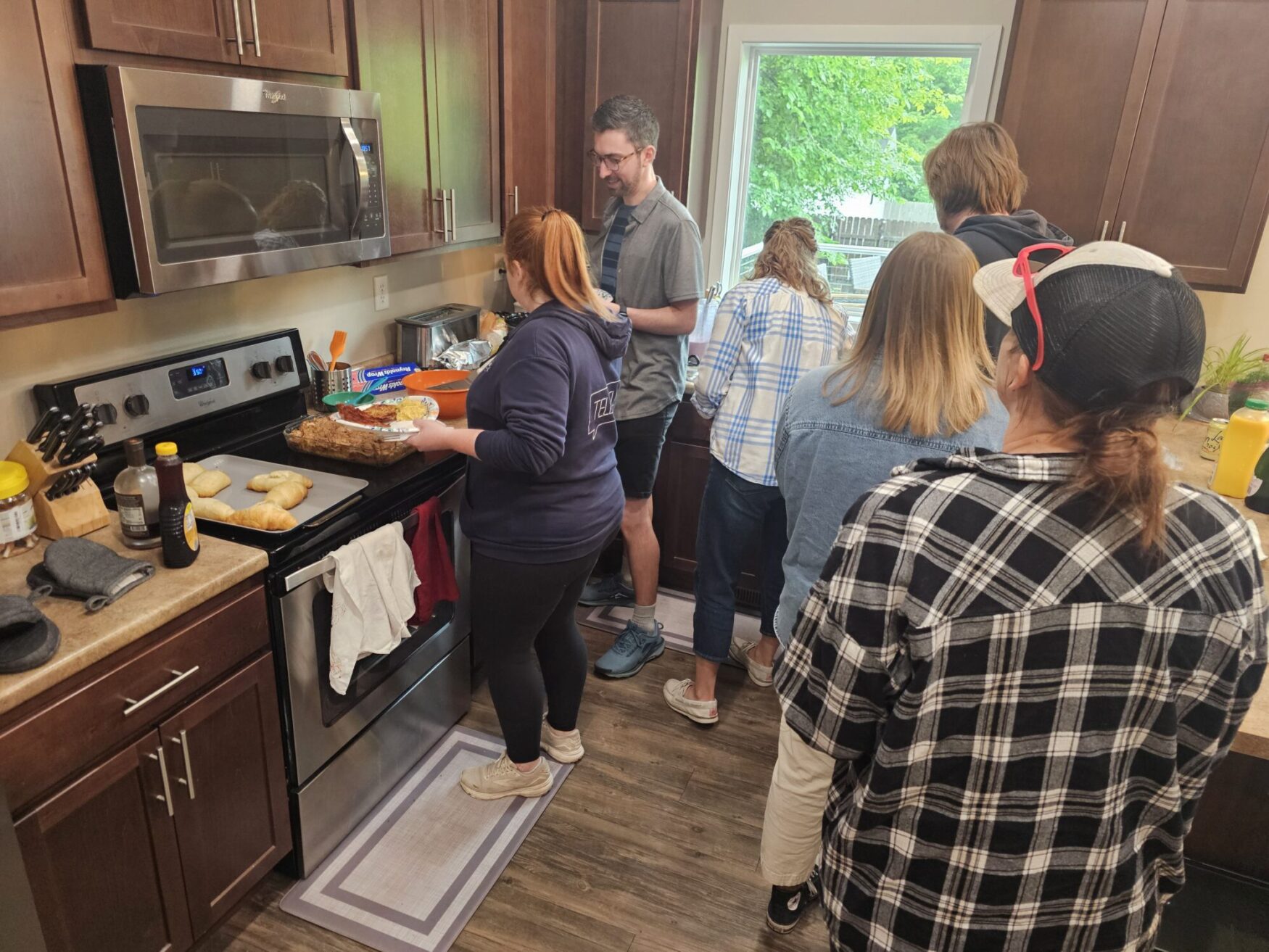 A group of people stand in line in a kitchen. They are each holding a plate, waiting to get breakfast.