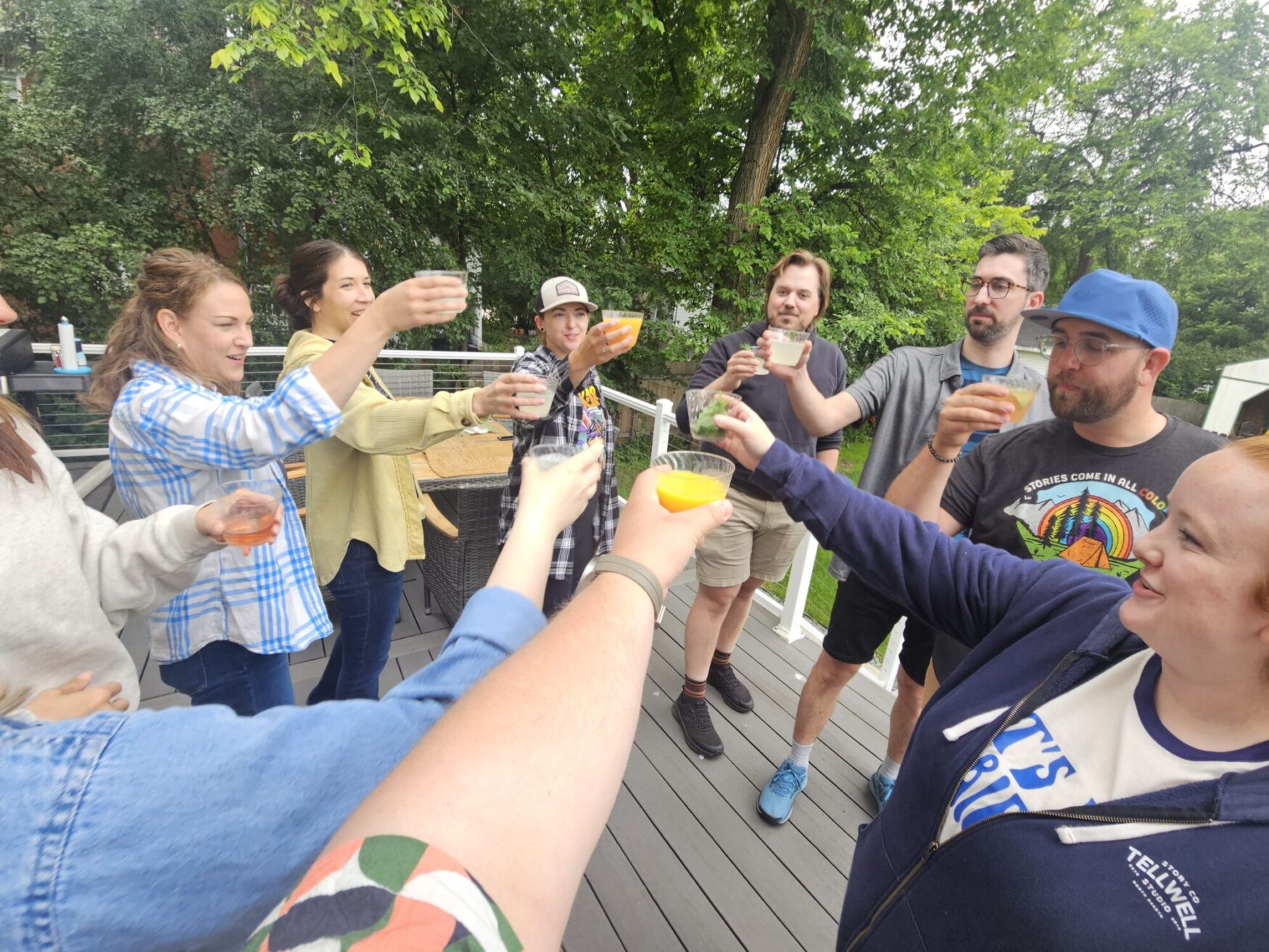 A group stands in a circle, each holding a glass giving a toast. They are outside standing on a deck surrounded by trees.