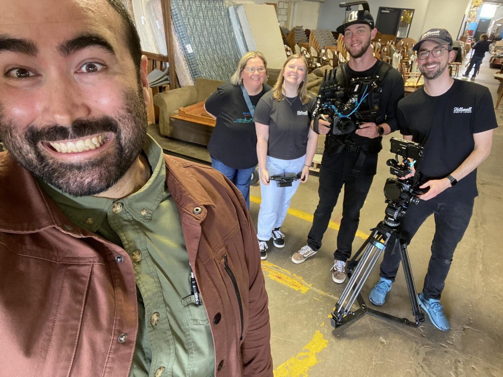 A group of five people pose for a cheerful photo inside a warehouse-style space filled with furniture. In the foreground, a man in a rust-colored jacket takes a close-up selfie, smiling widely. Behind him, two women and two men—some holding video equipment and wearing "Tellwell" branded shirts—stand together smiling. One man is wearing a camera rig, and another is standing behind a tripod-mounted camera. The setting suggests a video shoot at a furniture donation or resale center.