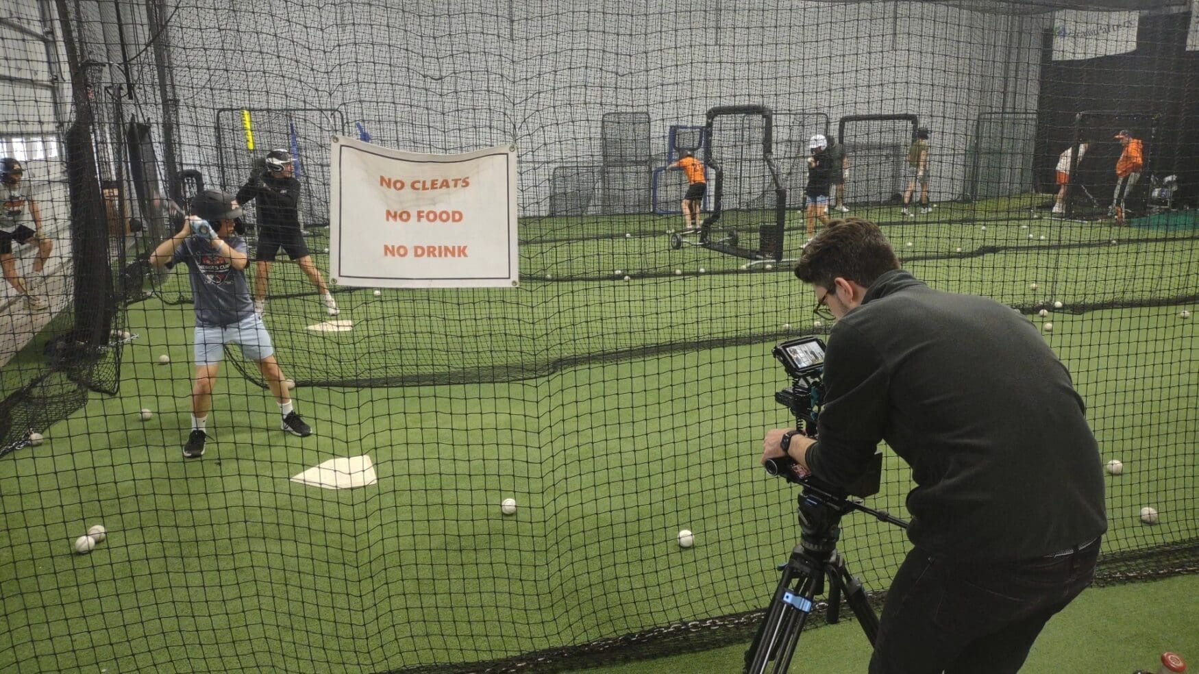 A filmmaker records a young baseball player preparing to swing inside an indoor training facility, with other players practicing in the background behind netting and a visible sign that reads "No cleats, no food, no drink."