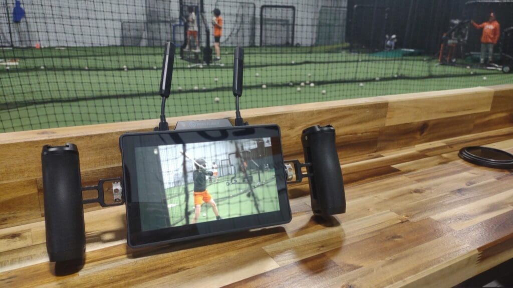 A wireless monitor on a wooden bench displays live footage of a young baseball player at bat inside an indoor training facility, while the practice continues in the background beyond a protective net.