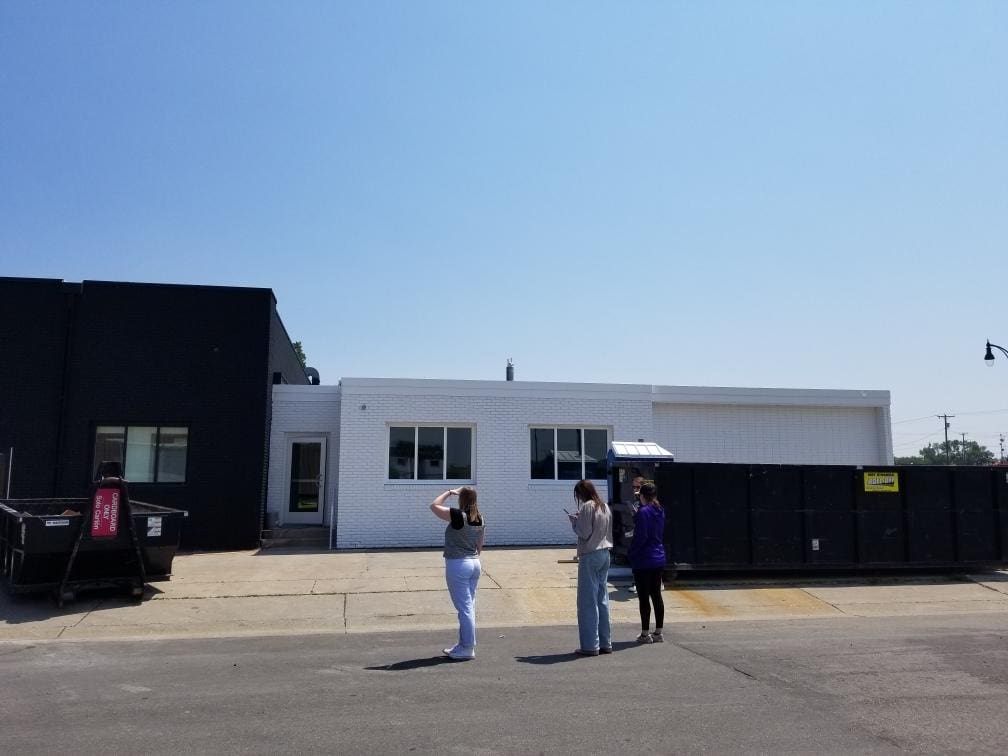 Three people stand on a sunny street in front of a white and black building under renovation, with large dumpsters positioned along the sidewalk.