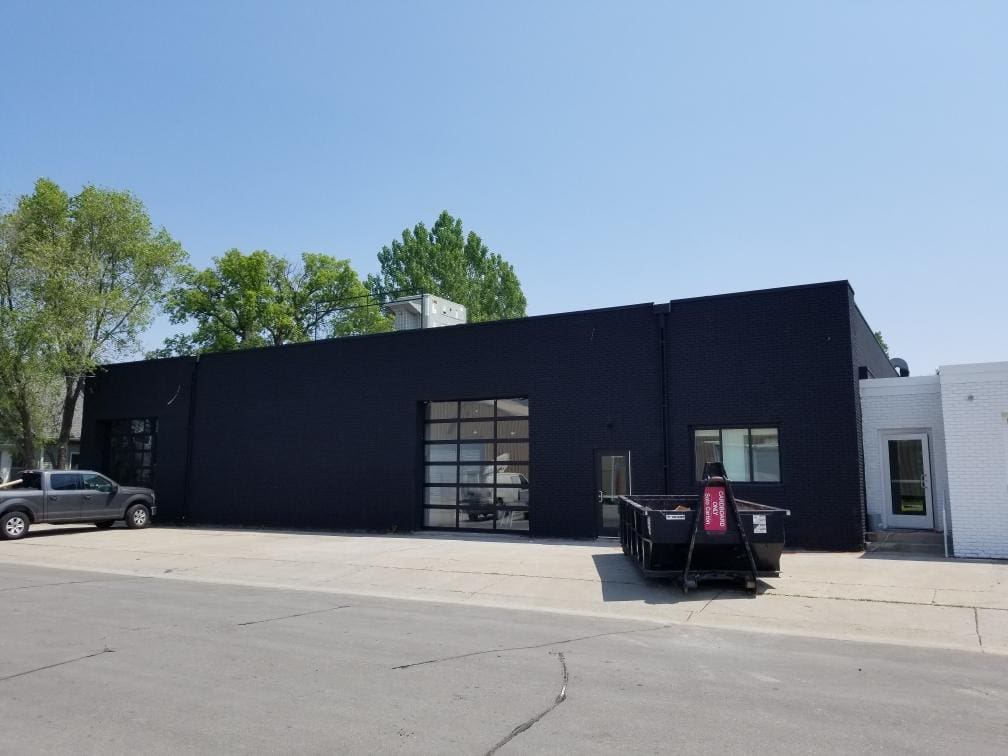 A black-painted commercial building with large windows and a roll-up garage door sits under a clear blue sky, with a parked pickup truck and construction dumpster out front.