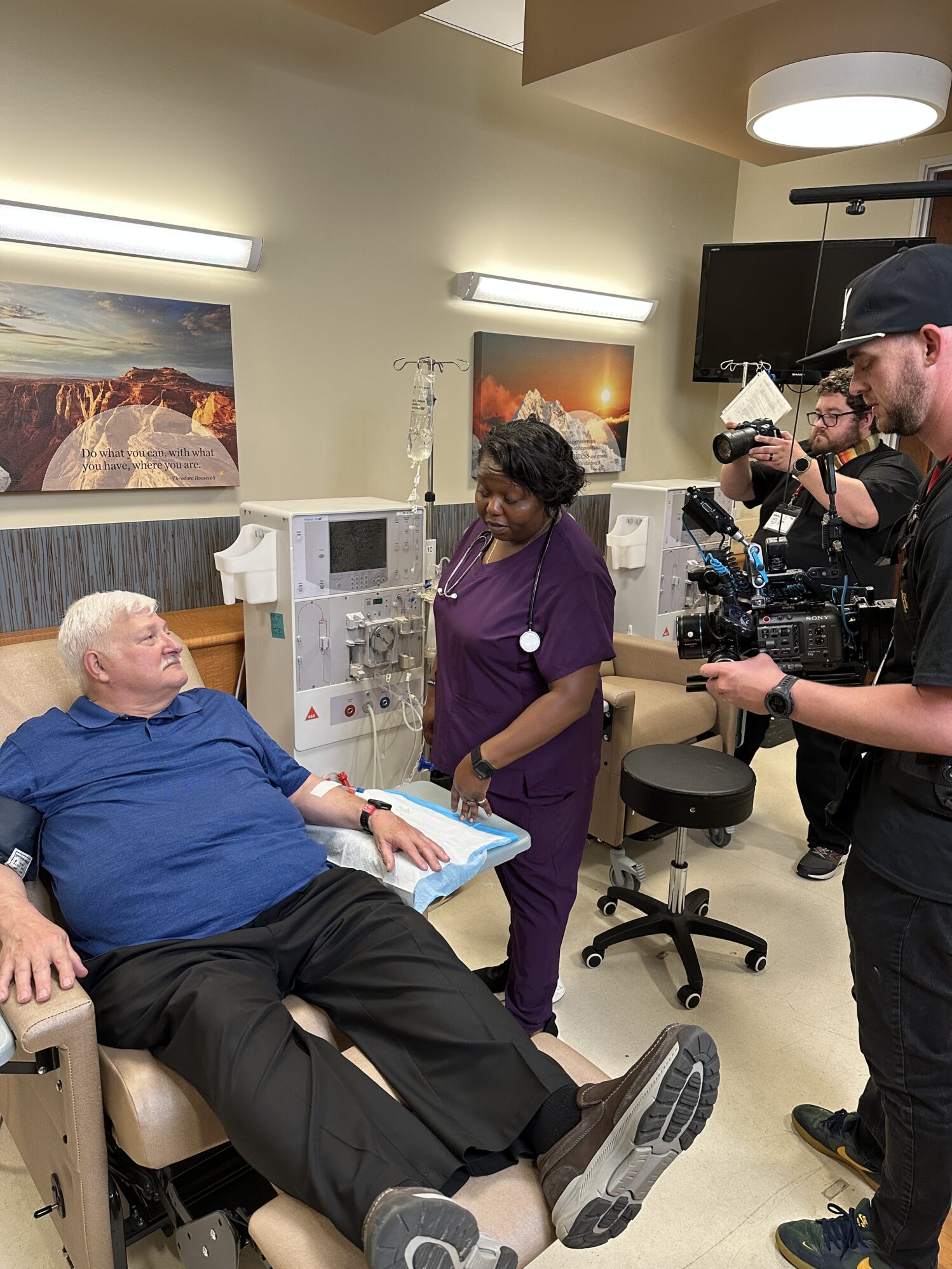 An older man sits in a kidney dialysis chair as a female nurse wearing purple scrubs gives him care. A man holds a video camera, filming the interaction. Another man stands next to the camera operator with a handheld camera, taking photos of the scene.