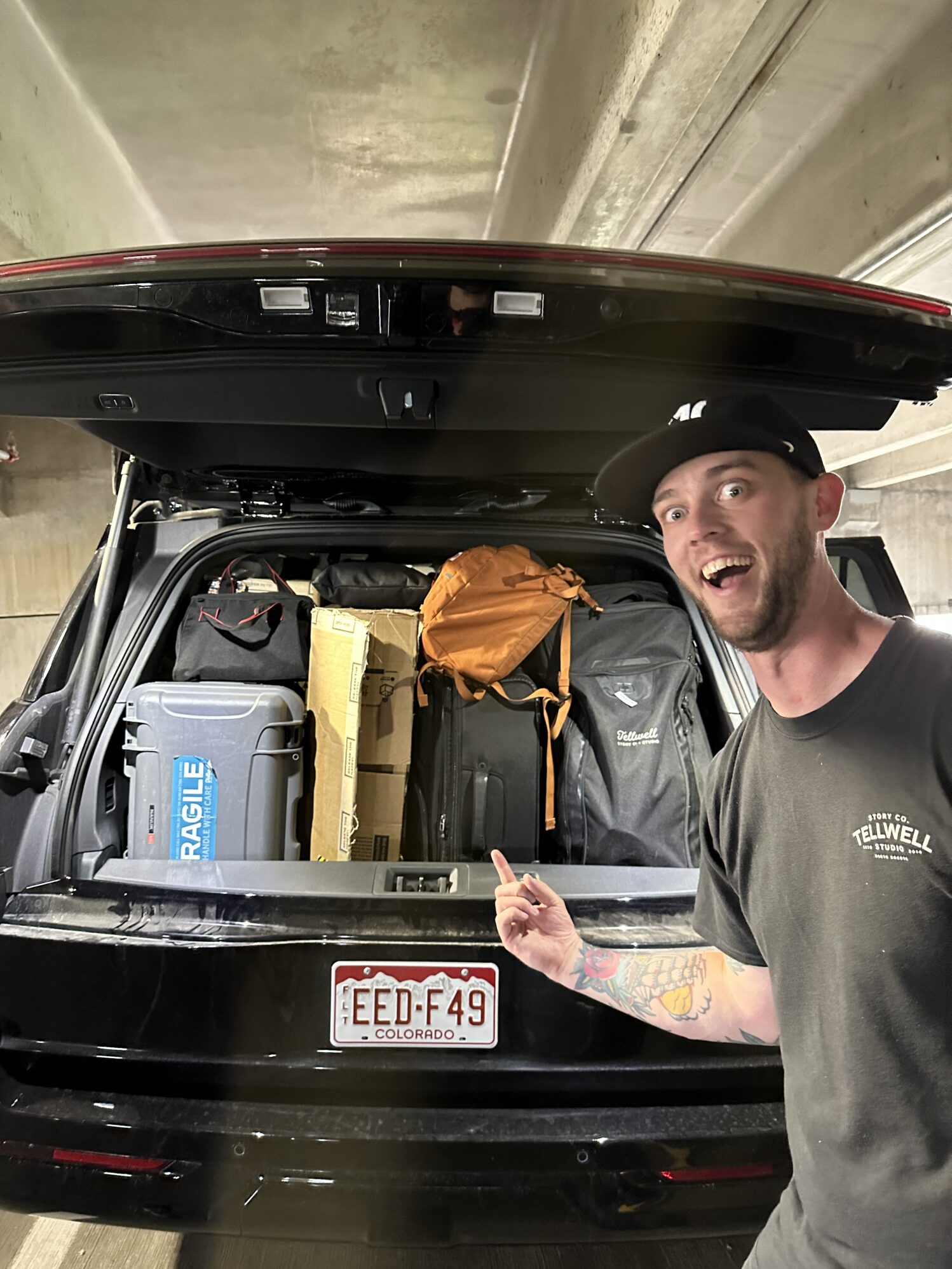 A man wearing a hat and a black shirt stands next to a car with the trunk door open, pointing at the full car filled with suitcases, film gear, and backpacks. His face is comical because the car is so full.