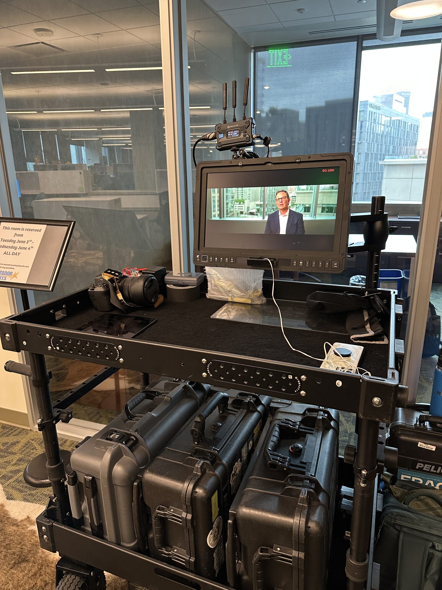 A two-layered utility cart filled with camera equipment. On the bottom shelf, there are three hard cases lined up neatly. On the top shelf, there is a tablet, a phone, and a camera. There is also a large monitor attached to the cart, showing a man in an interview setting.