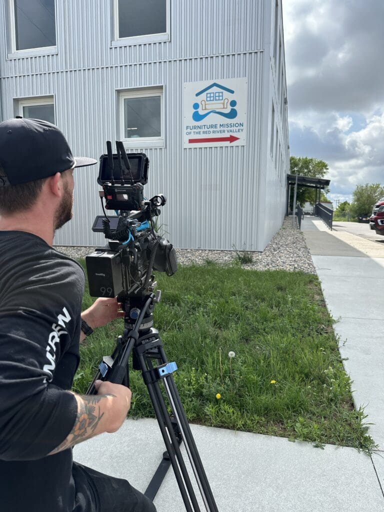 A man operating a professional video camera on a tripod is filming outside a gray metal building. The building has several windows and a sign that reads "Furniture Mission of the Red River Valley" with a directional arrow. The scene takes place on a partly cloudy day, and there is a patch of grass and a paved walkway in the foreground.