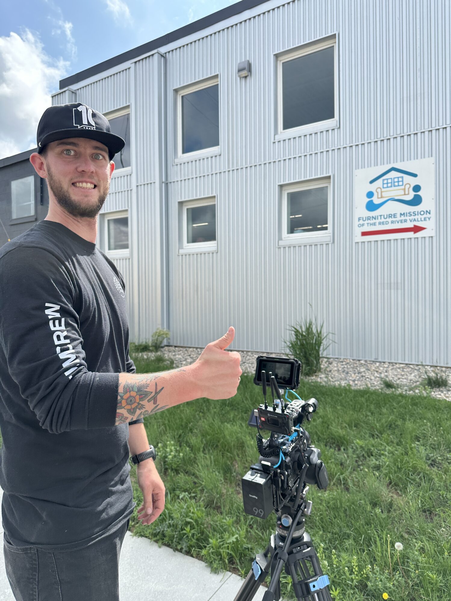 A man operating a professional video camera on a tripod is filming outside a gray metal building. He is smiling at the camera and giving a thumbs up. The building has several windows and a sign that reads "Furniture Mission of the Red River Valley" with a directional arrow. The scene takes place on a partly cloudy day, and there is a patch of grass and a paved walkway in the foreground.