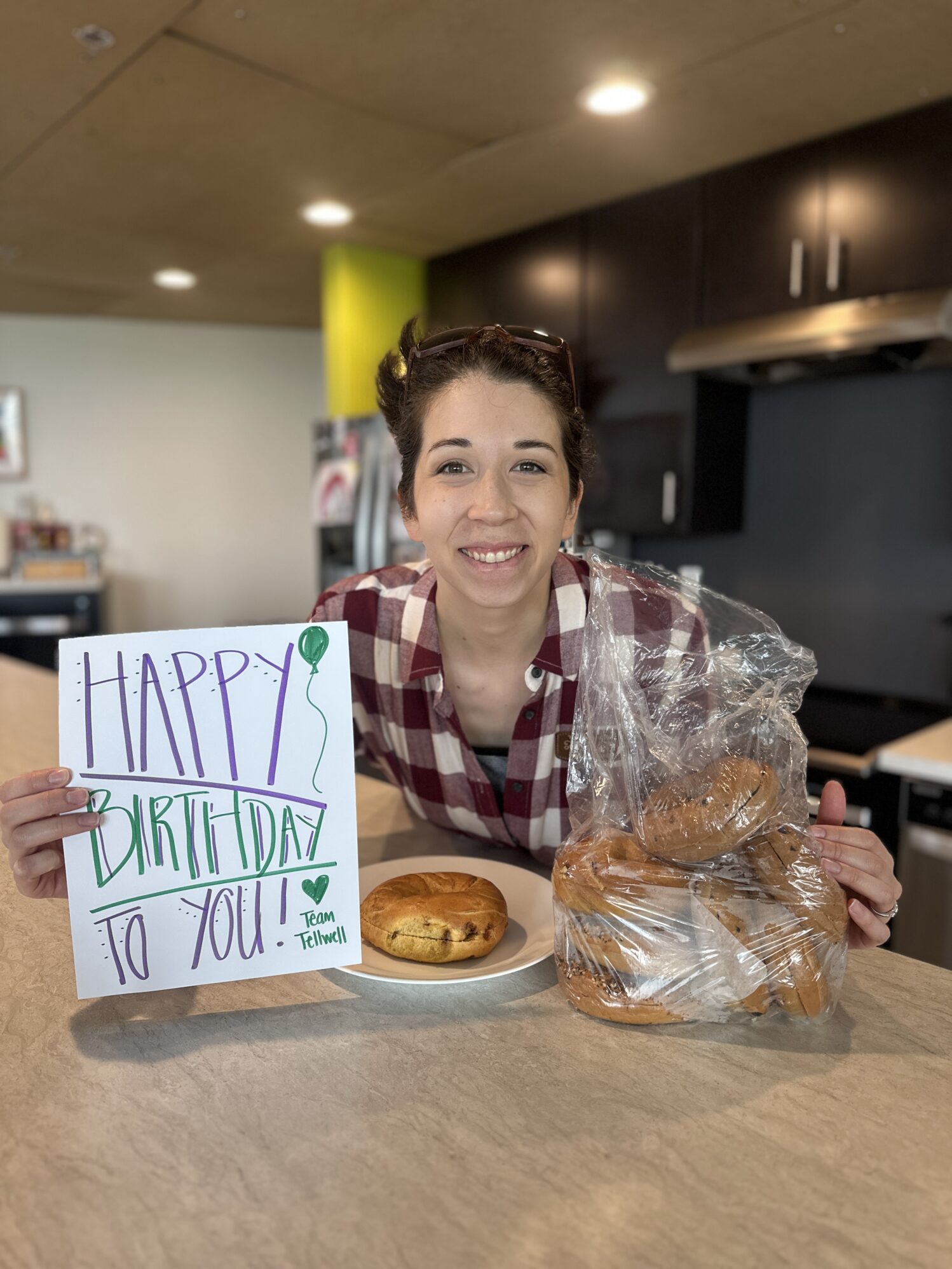 A smiling woman in a red plaid shirt poses in a kitchen area, holding a colorful sign that reads "HAPPY BIRTHDAY TO YOU! ♥ Team Tellwell" with a green balloon drawing. In front of her is a plate with a single bagel and a large plastic bag filled with more bagels. The background features modern dark cabinets and a stainless steel stove, with warm overhead lighting.