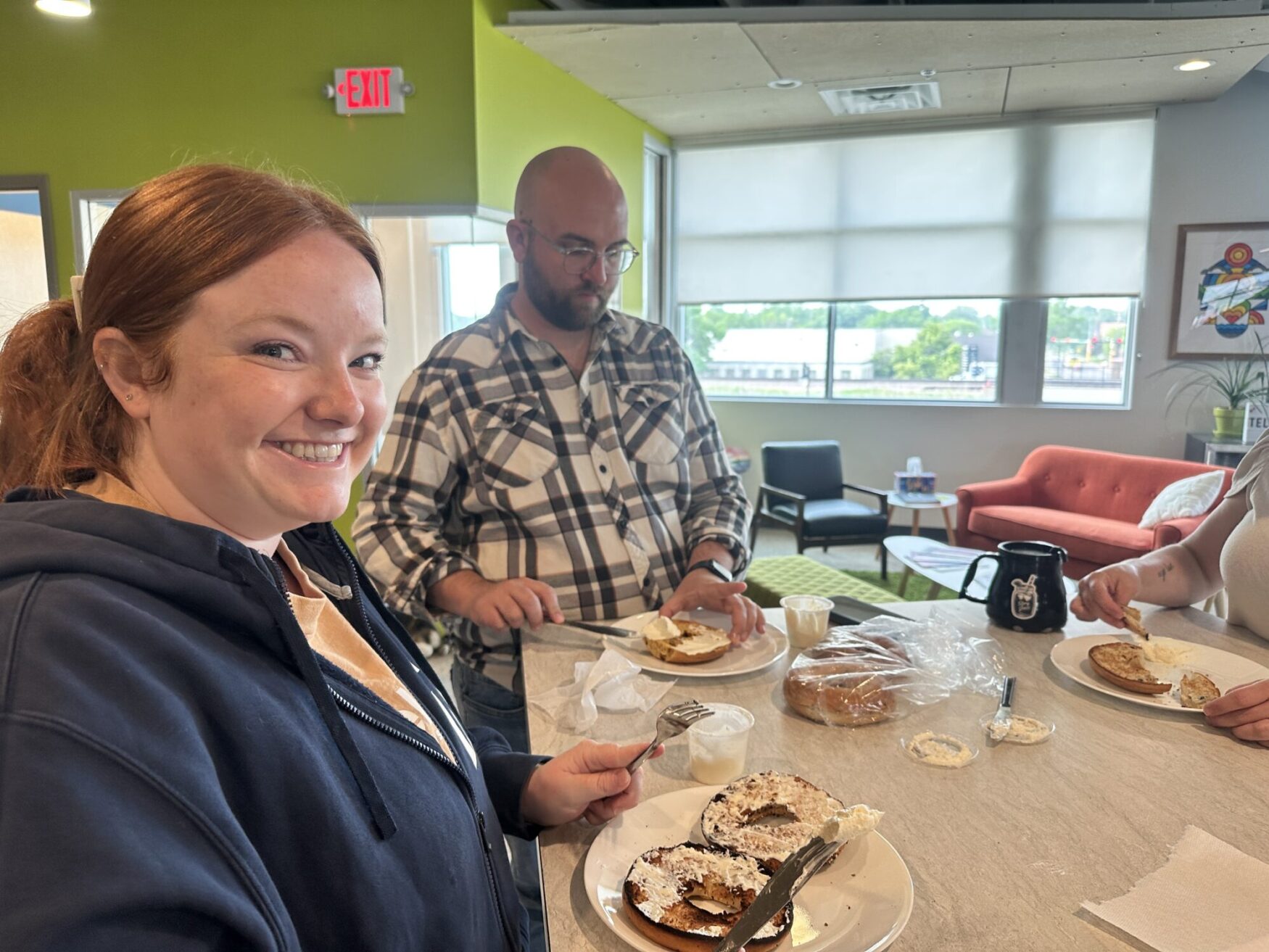 A cheerful group of people is gathered around a counter in a brightly lit office kitchen, preparing bagels with cream cheese. A smiling woman in the foreground wears a dark hoodie, holding a plate with a toasted bagel. Behind her, a man in a plaid shirt is focused on spreading cream cheese. Another person’s arms are visible on the right, also making a bagel. A black coffee mug and a bag of bagels are on the counter. The background features large windows, a red couch, and colorful wall art.