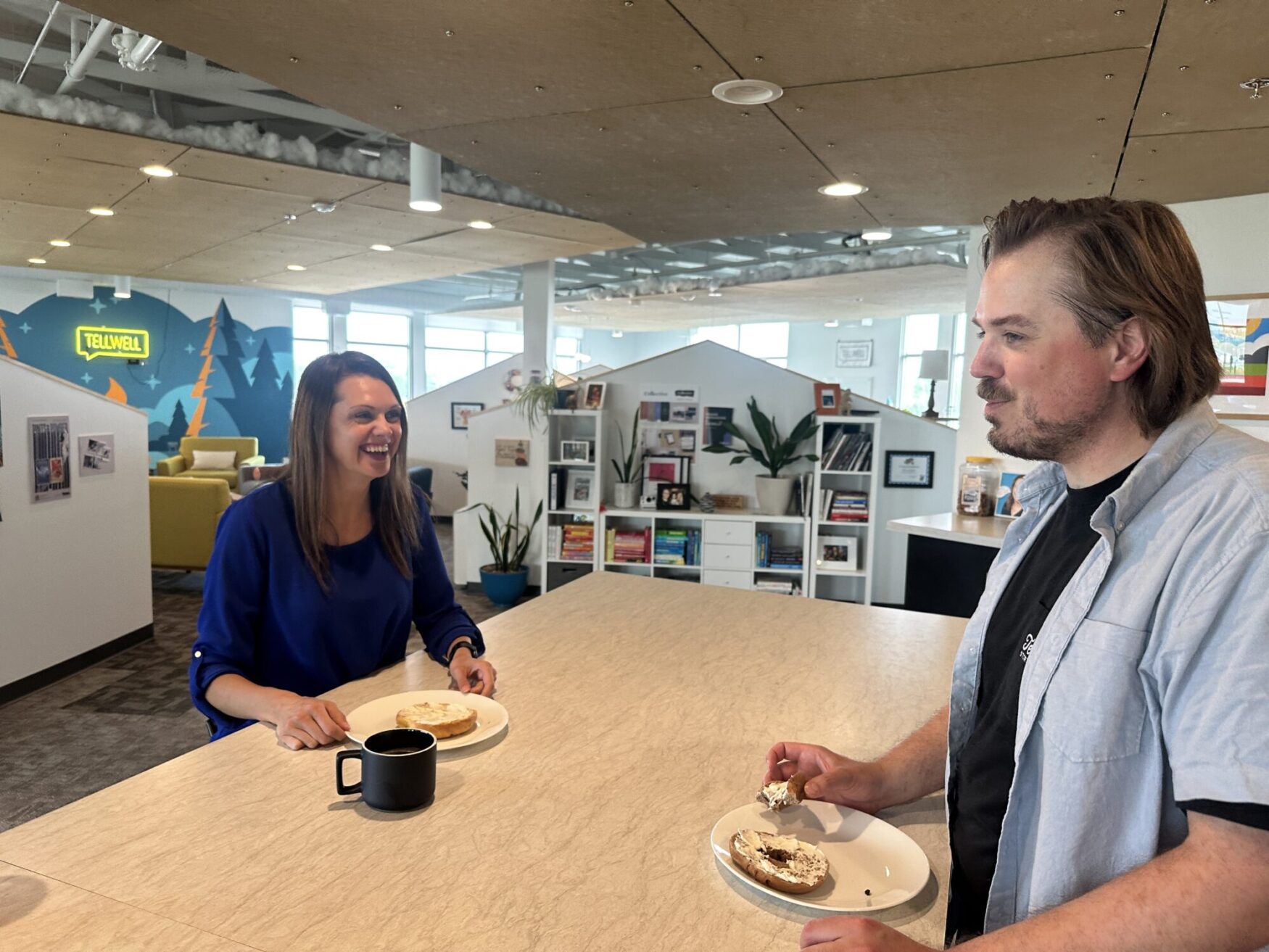 Two people are enjoying a casual conversation while eating bagels in a modern office kitchen area. The woman on the left is smiling and wearing a royal blue blouse, while the man on the right, dressed in a black shirt and light blue overshirt, is mid-sentence. Both have plates with bagels and coffee mugs in front of them. The background includes bright decor, a colorful mural with the word "TELLWELL," bookshelves, and plants.