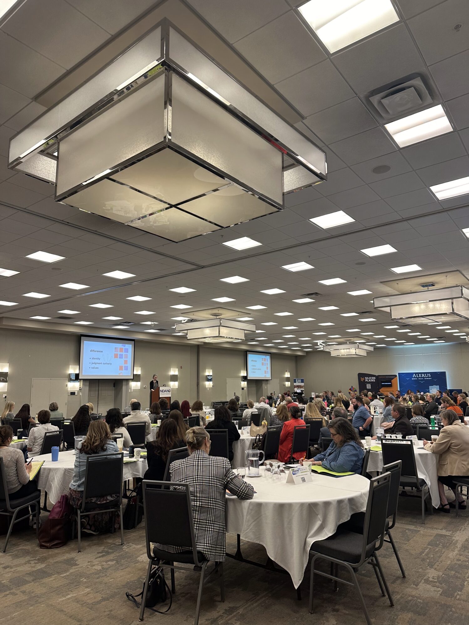 A large conference room filled with round tables and attendees listening to a speaker at a podium. Two projection screens display a slide titled “differences” with a colorful grid. Overhead lighting is modern and geometric, and sponsor banners including Alerus and The Title Team are visible in the back of the room.