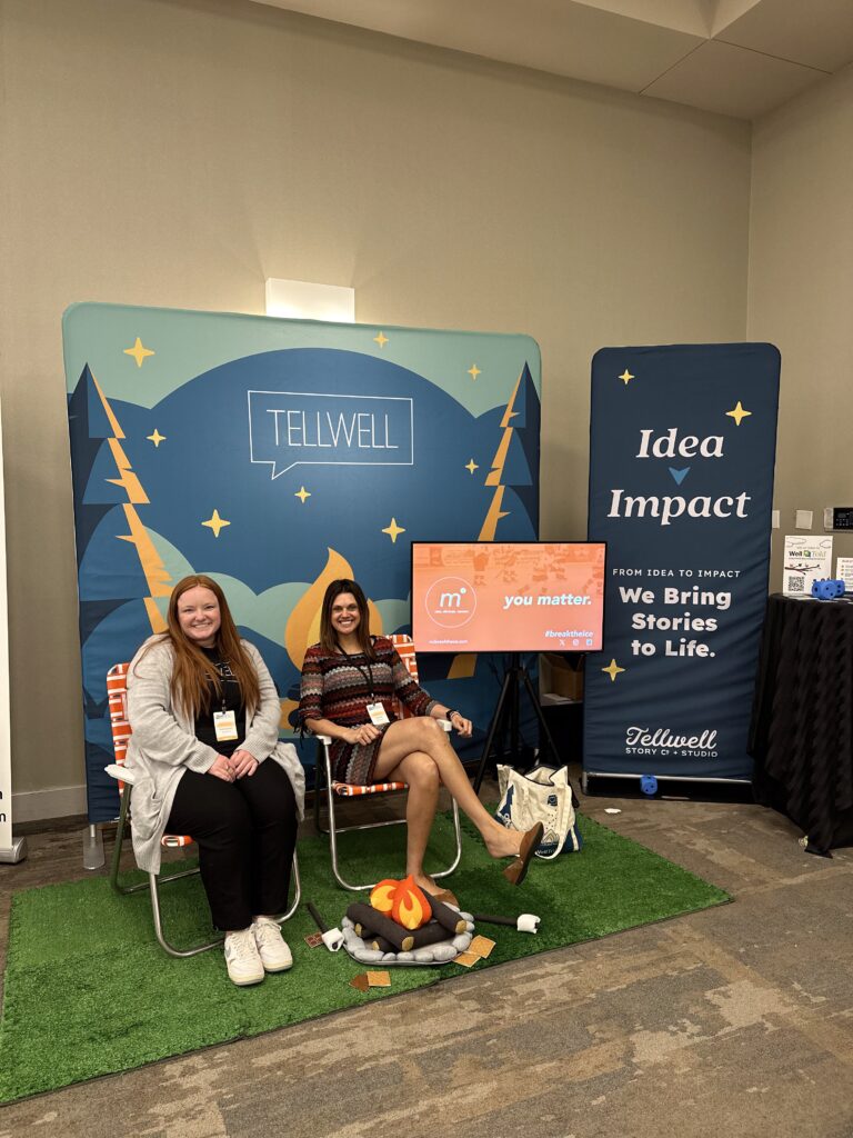 Two women sit in orange camp chairs on a small patch of artificial grass, in front of a colorful Tellwell-branded backdrop featuring trees, stars, and a campfire scene. Between them is a display of plush campfire props, including a felt fire, marshmallows, and graham crackers. Behind them, a sign reads “Idea → Impact” and “We Bring Stories to Life,” along with Tellwell’s logo. A monitor beside them displays the message “you matter.” with hashtags and a link to #breaktheice.