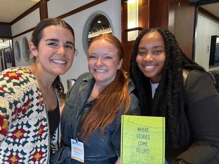 Three women smile at the camera. The woman in the middle is wearing a lanyard with a name tag at an event.