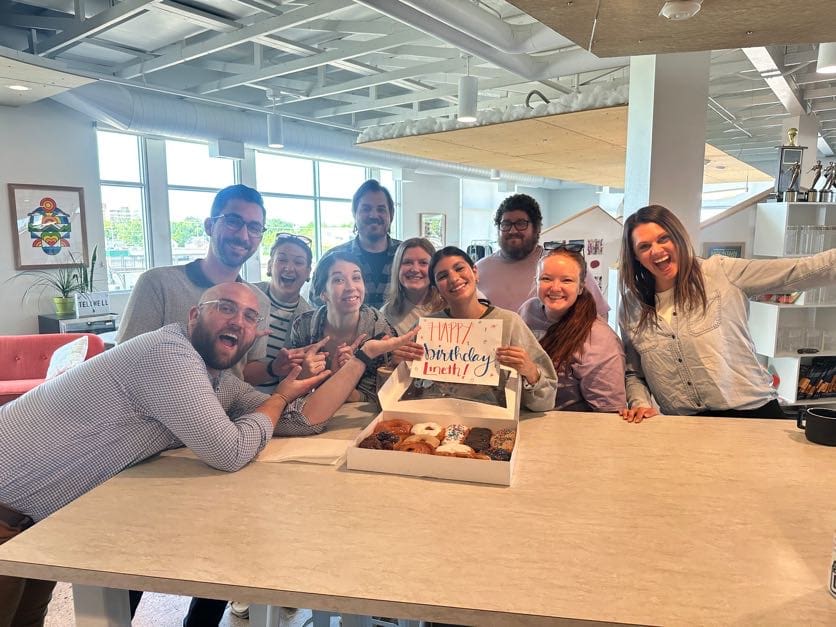 A group of eleven coworkers gathered around a table in a bright, modern office, celebrating a birthday. A woman in the center holds a sign that reads “Happy Birthday Lineth!” with an open box of assorted donuts in front of her. Everyone is smiling and posing enthusiastically, creating a cheerful and festive atmosphere.