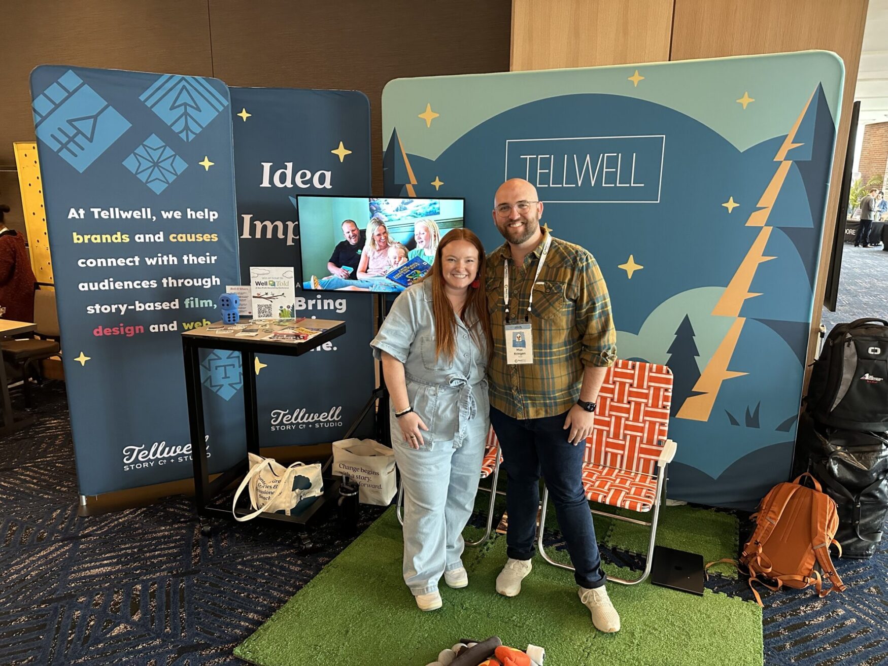 Two people smile in front of a colorful Tellwell Story Co. booth at a conference, featuring branded signage, a TV displaying video content, promotional items, and playful décor including a turf mat and lawn chair.