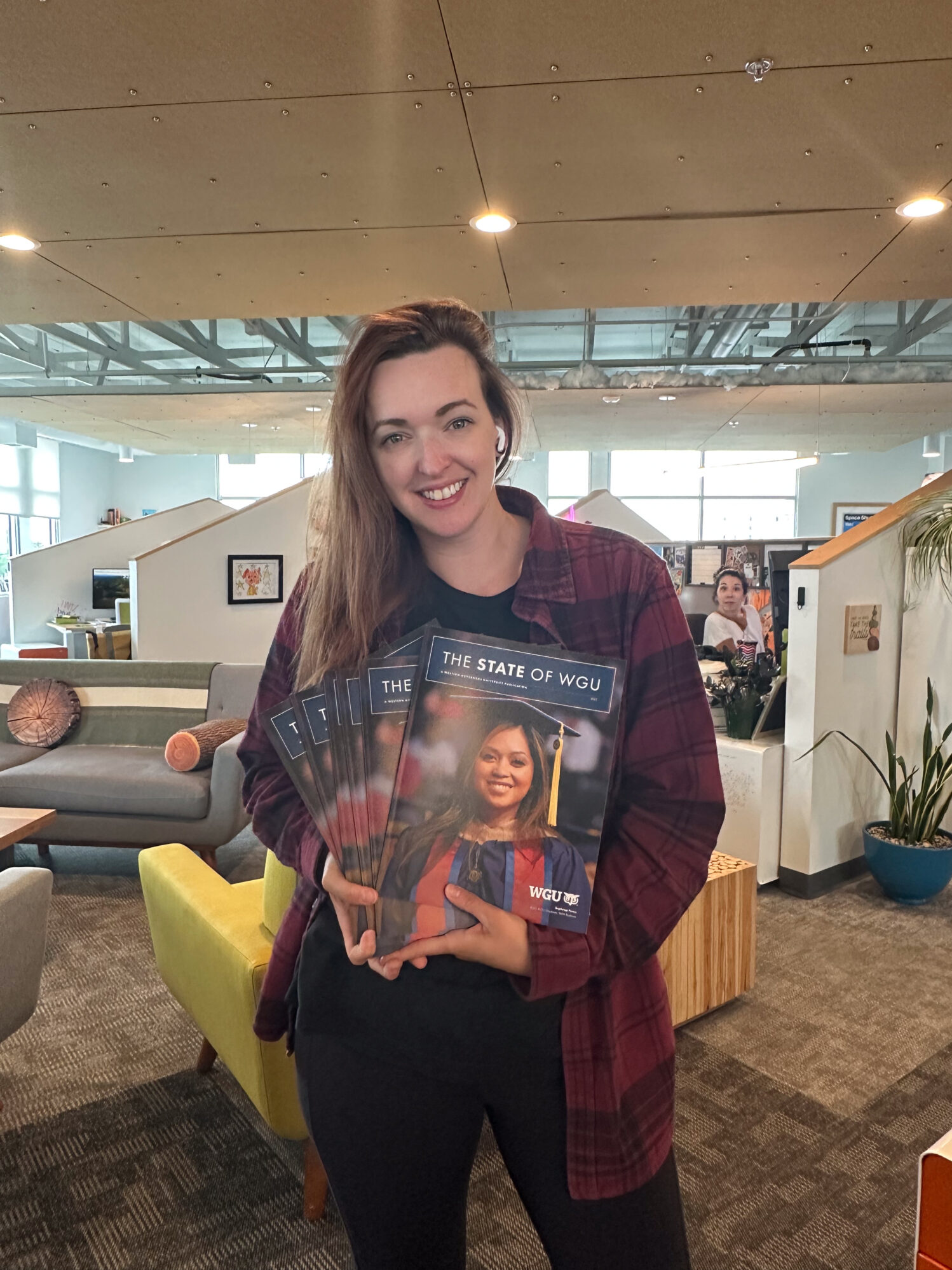 A woman in a long sleeve, plaid shirt, holding a stack of magazines in an office setting.