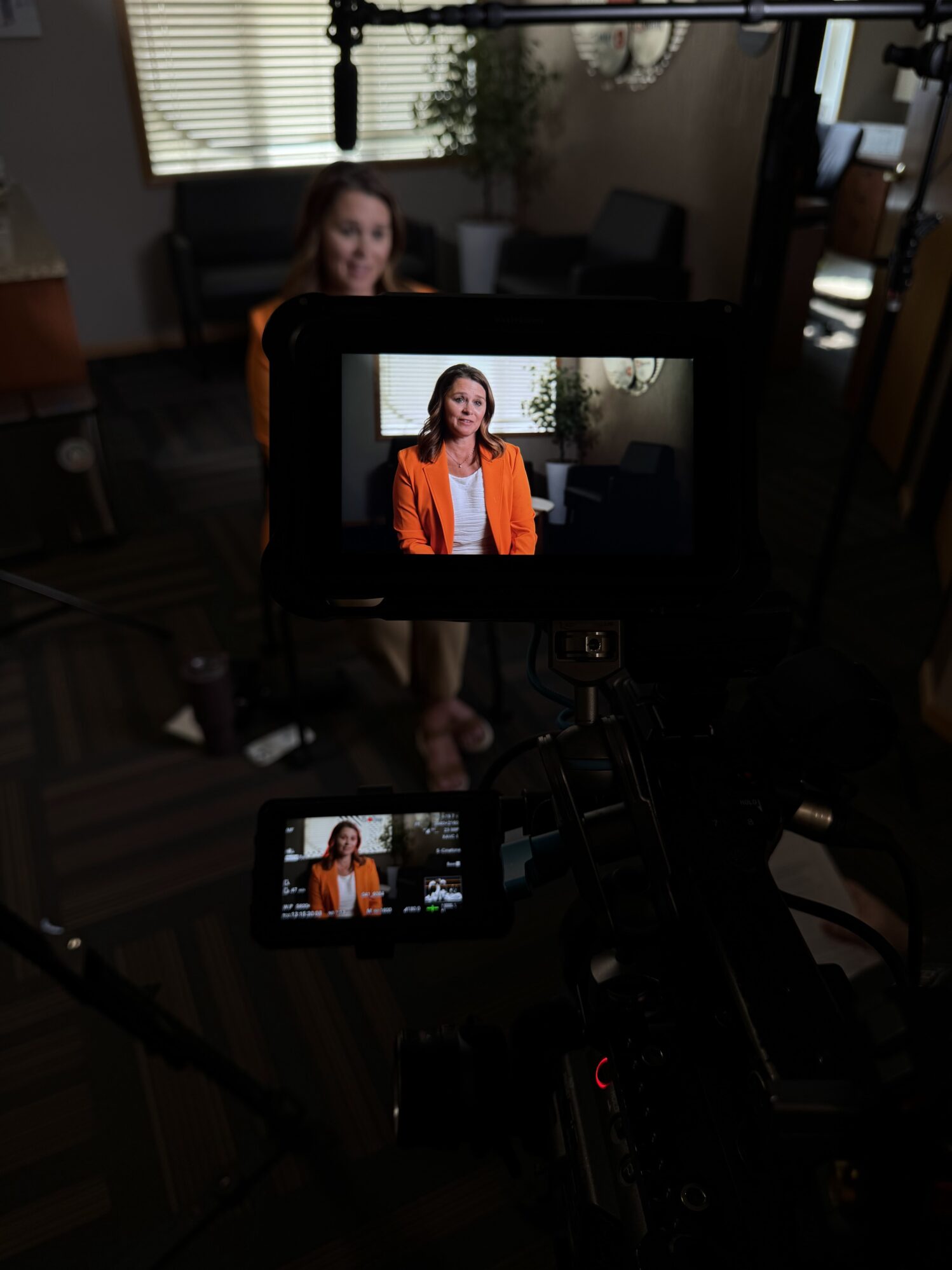 A woman in an orange blazer is framed on a camera monitor during a professional video interview setup, with lights, microphones, and crew equipment partially visible in the dimly lit room.