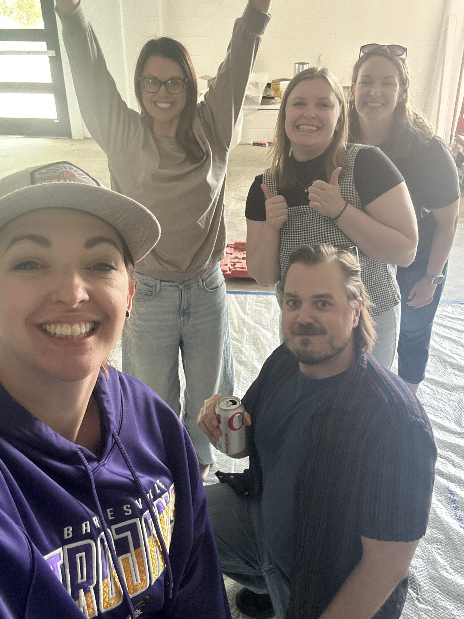 A group of 4 women and one man pose for a selfie. They look excited and happy. One woman is holding her arms straight up, while another is giving two thumbs up. The man is kneeling on the ground holding a can of Diet Coke.