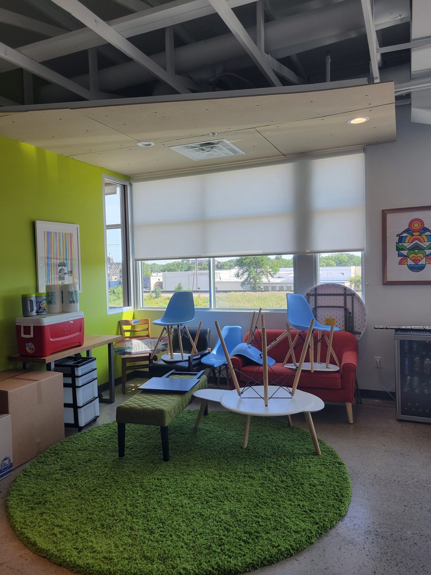 A colorful lounge area in an office space with a lime green accent wall and large windows letting in natural light. Furniture is stacked as if the room is being rearranged or cleaned—blue chairs are placed upside down on a red sofa and white coffee tables. A green circular rug and various art pieces give the room a playful, casual vibe.