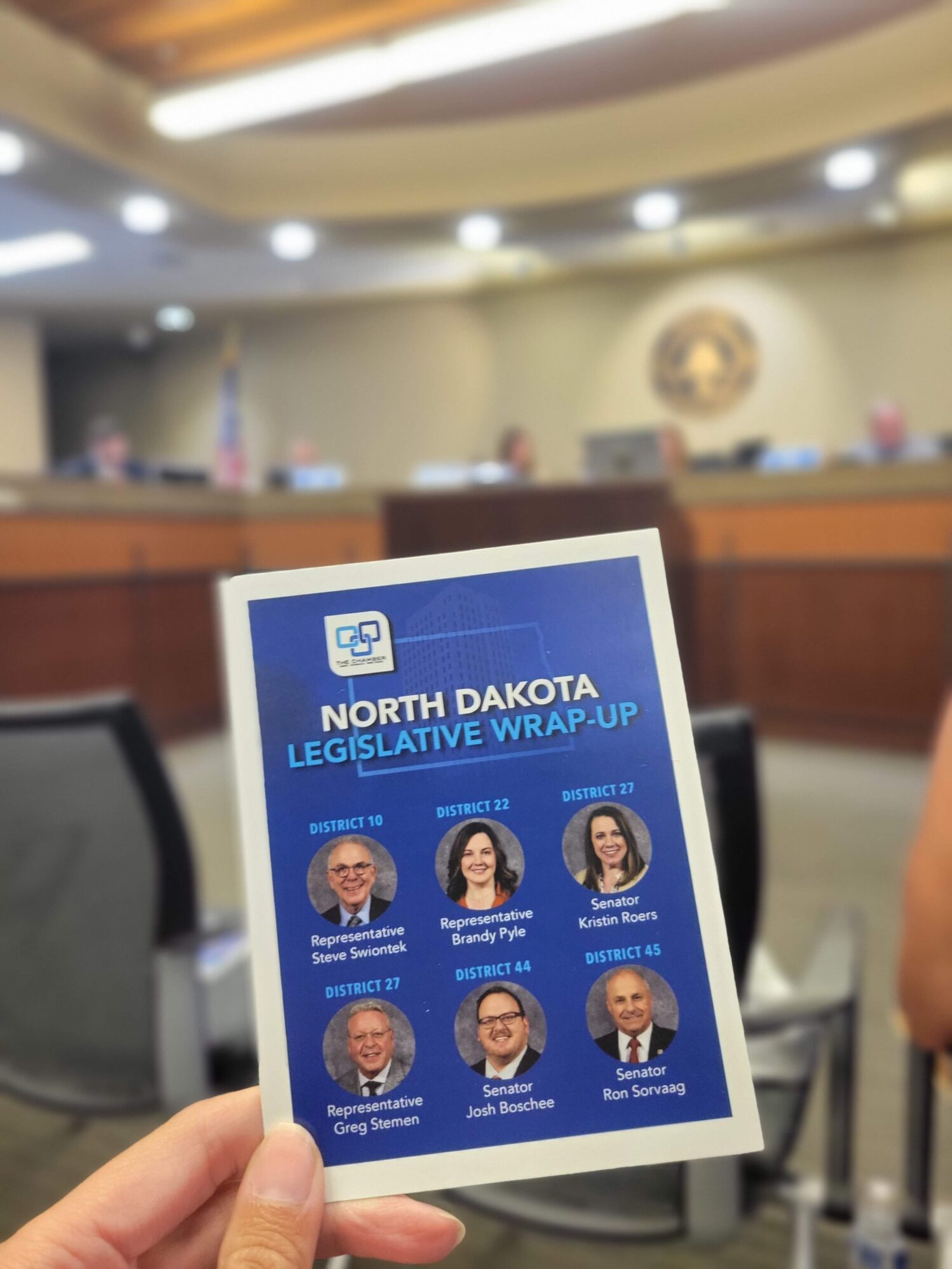 A person holds a pamphlet titled “North Dakota Legislative Wrap-Up” in the foreground of a government meeting room. The pamphlet features headshots and names of six state legislators, including Representatives Steve Swiontek, Brandy Pyle, Greg Stemen, and Senators Kristin Roers, Josh Boschee, and Ron Sorvaag, along with their respective districts. The background shows a blurred image of a panel of officials seated at a raised dais, with the American flag and a seal on the wall behind them. The setting suggests a public legislative or community meeting.