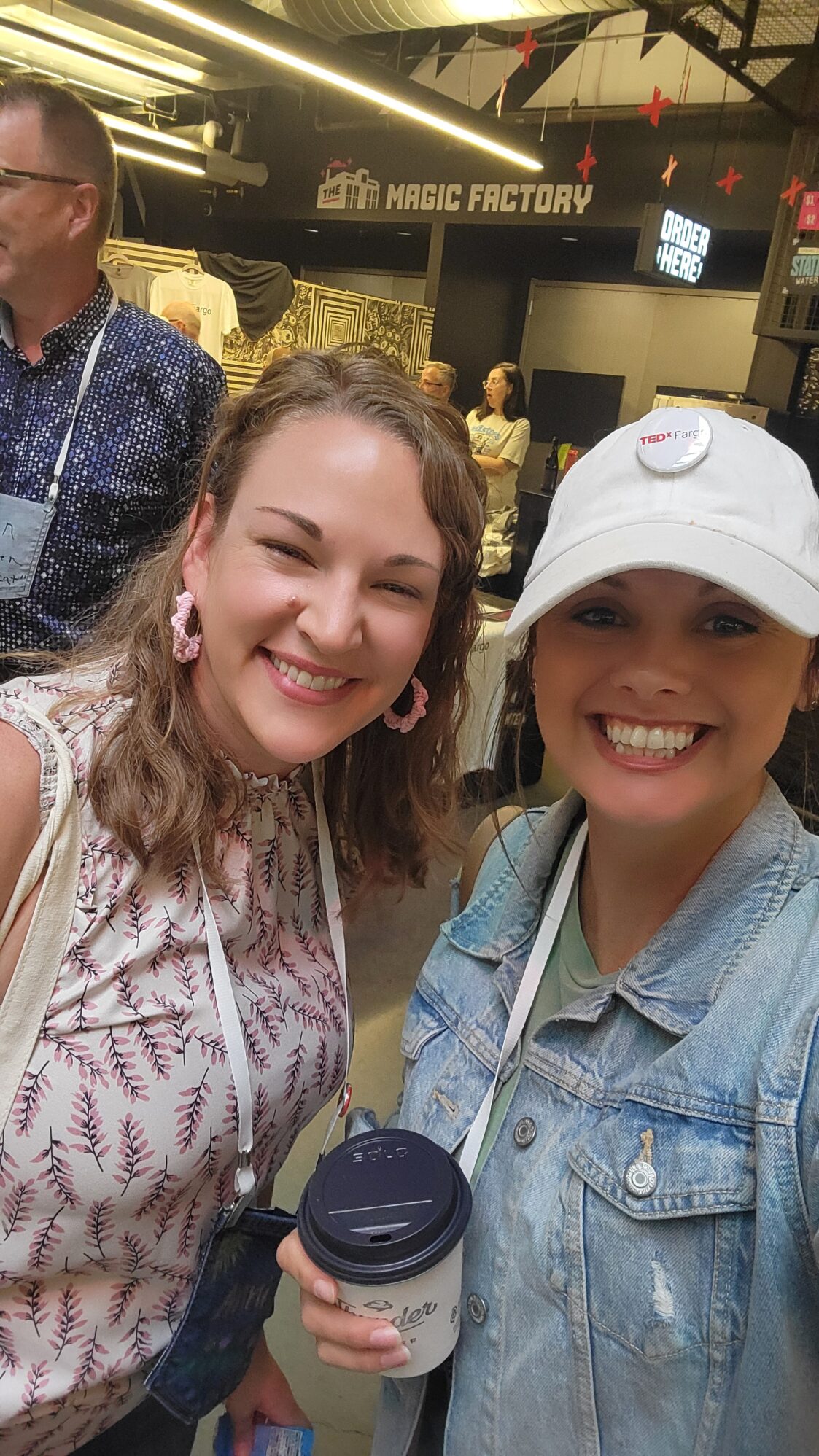 Two women smile for a selfie together. They are both wearing lanyards, suggesting they are at a corporate event. One woman wears a pin on her hat that reads “TEDxFargo” while she holds a cup of coffee. They are in an industrial looking room, with the words “Magic Factory" on the wall behind them.