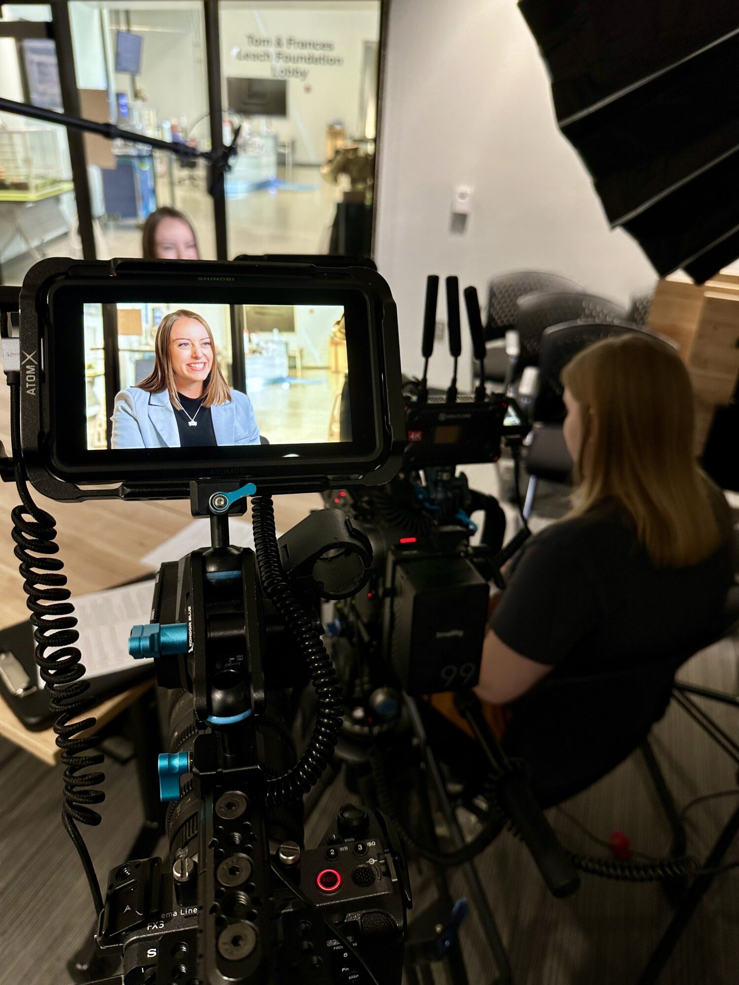 A blonde woman sits in a chair across from another woman who is being interviewed. There is a large camera with a screen showing the interviewee smiling.