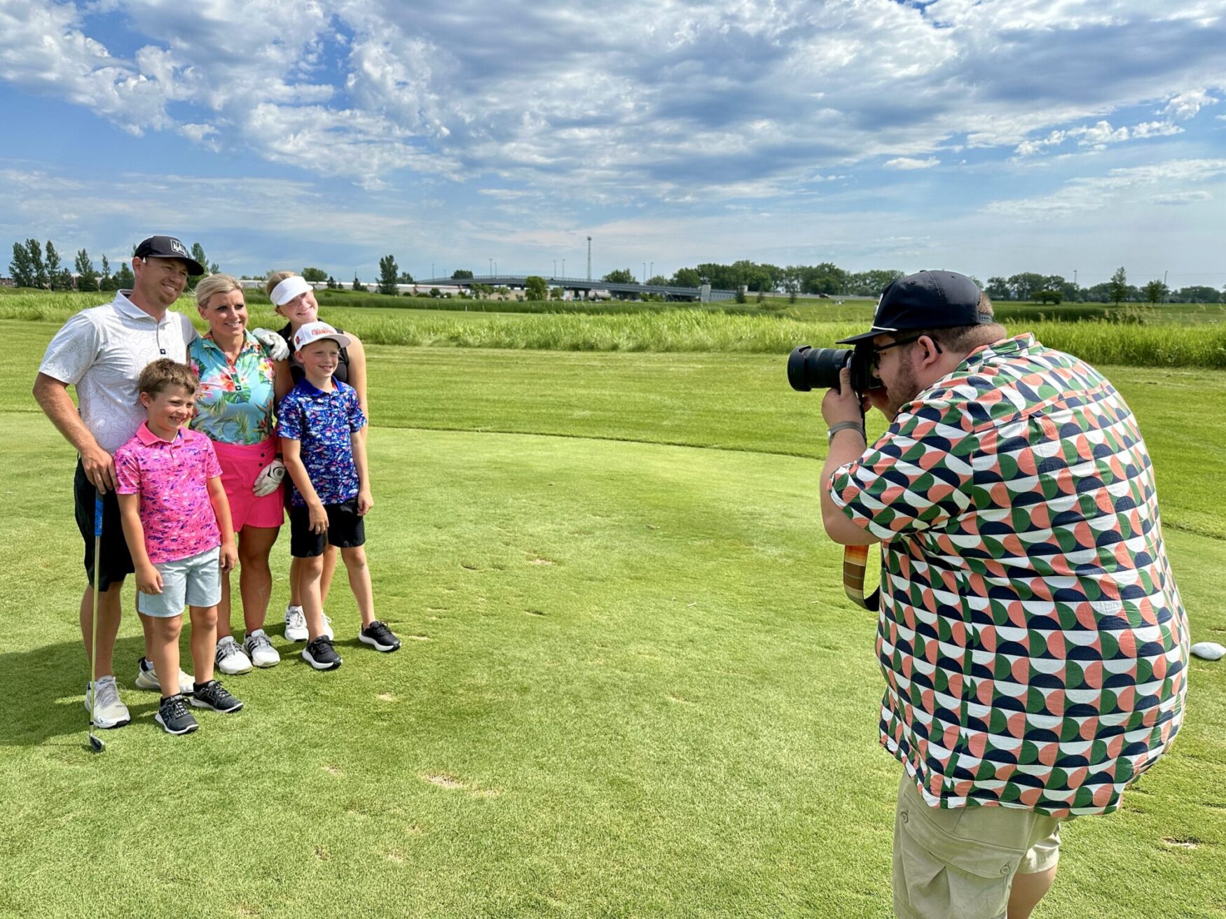 A man with a patterned shirt and black baseball cap holds a camera, taking a photo of a family of five. The family is golfing and standing on the green.