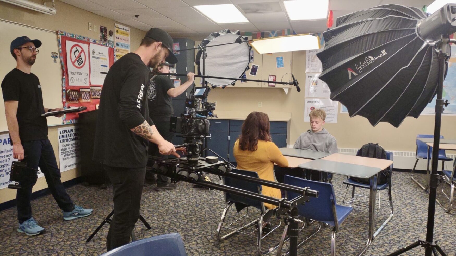 A film crew is set up in a classroom with a camera, lights, and audio. There are two actors sitting in school desks for this scene.