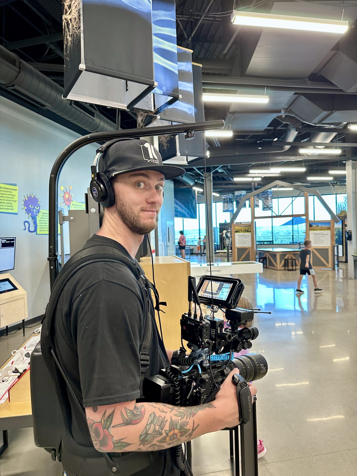 A man wearing a black shirt and black hat holds a large camera rig. He is smirking in the photo while holding the camera. He appears to be in a science museum.