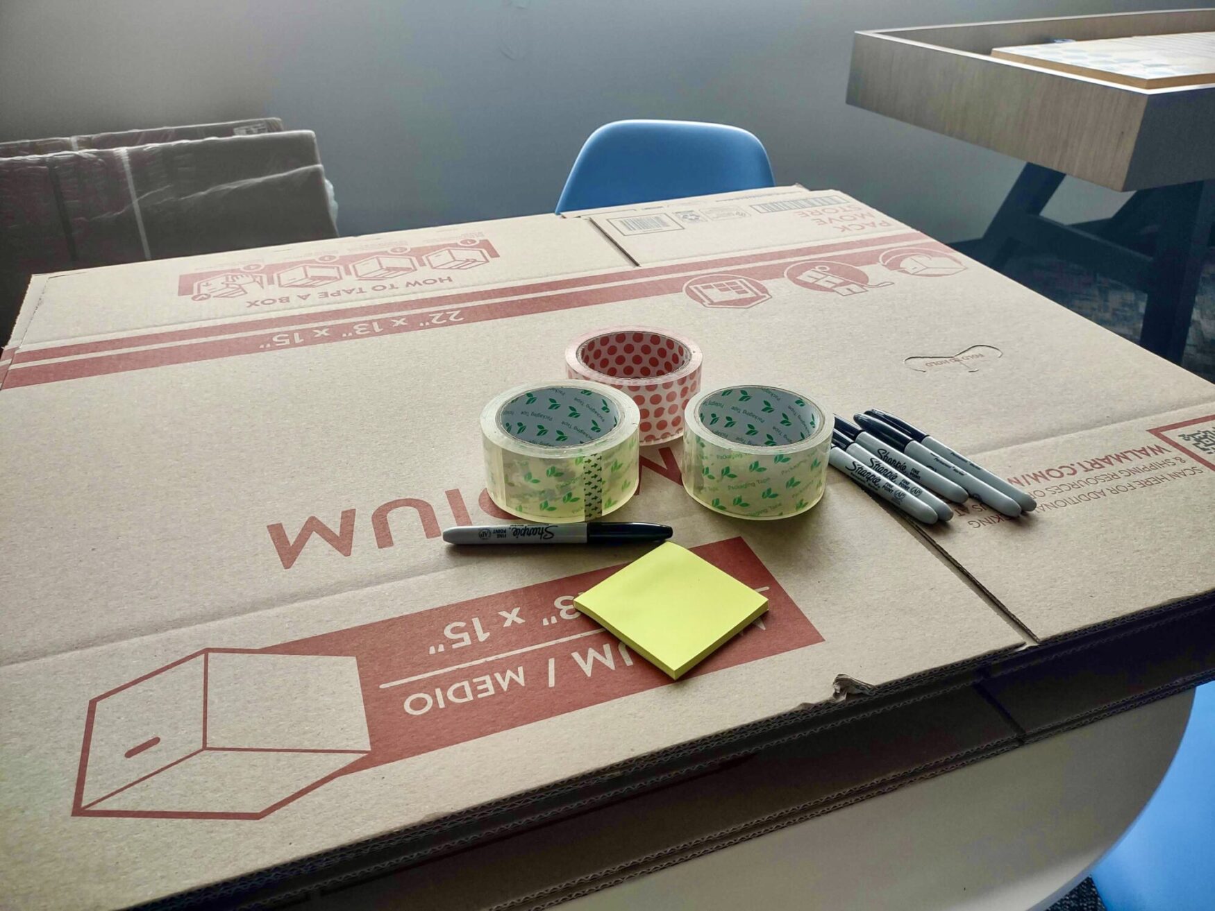 A stack of flattened moving boxes sits on a table along with three rolls of packing tape, several permanent markers, and a stack of StickyNotes.