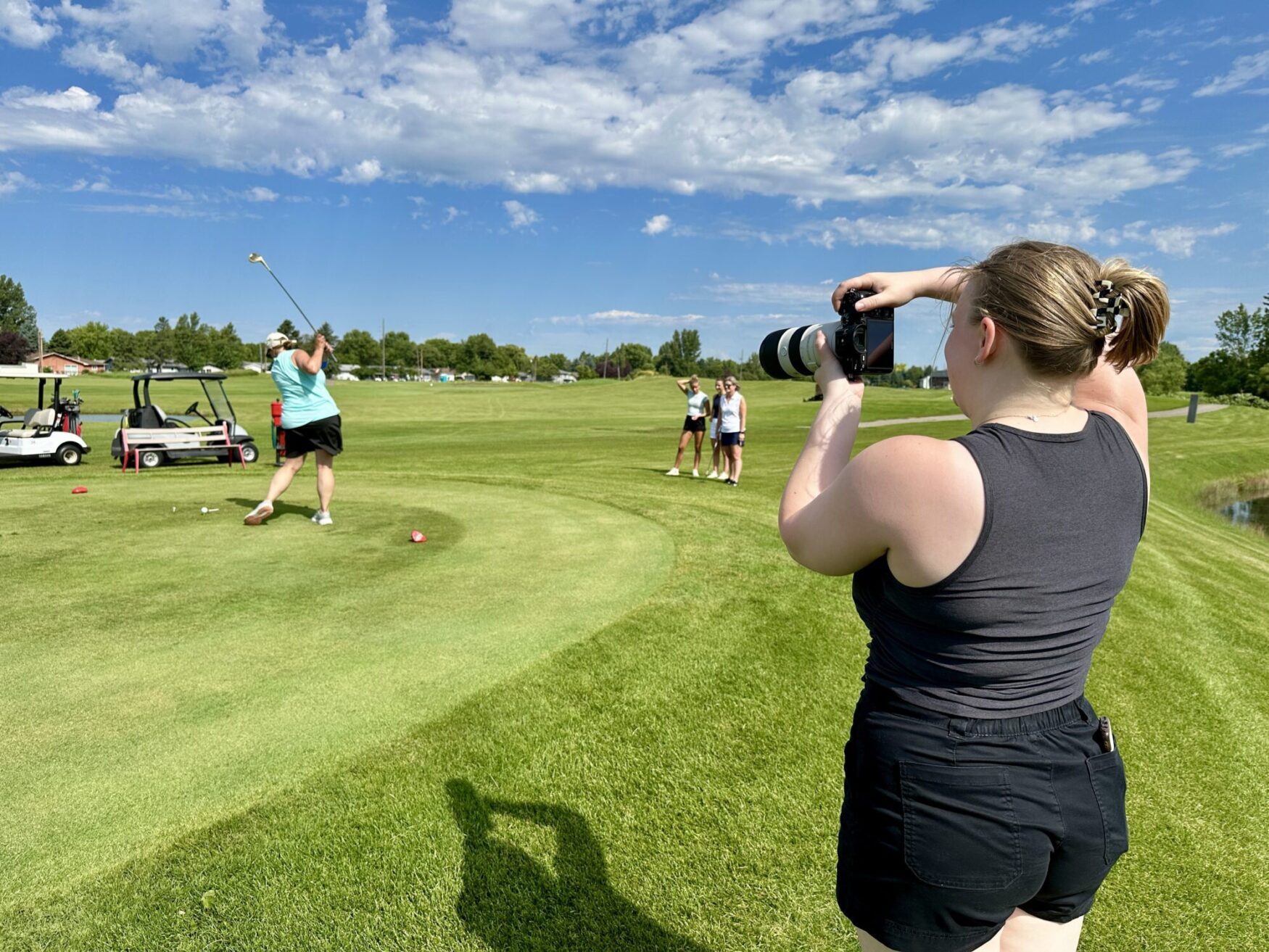 A woman in a black tank top and black shorts is holding a professional camera, photographing a golfer mid-swing on a sunny golf course. The golfer, wearing a light blue tank top and black skirt, is finishing a drive on the tee box. In the background, three women pose for a photo on the fairway, smiling. Golf carts and equipment are visible nearby, and the sky is bright with scattered clouds over the lush green course.