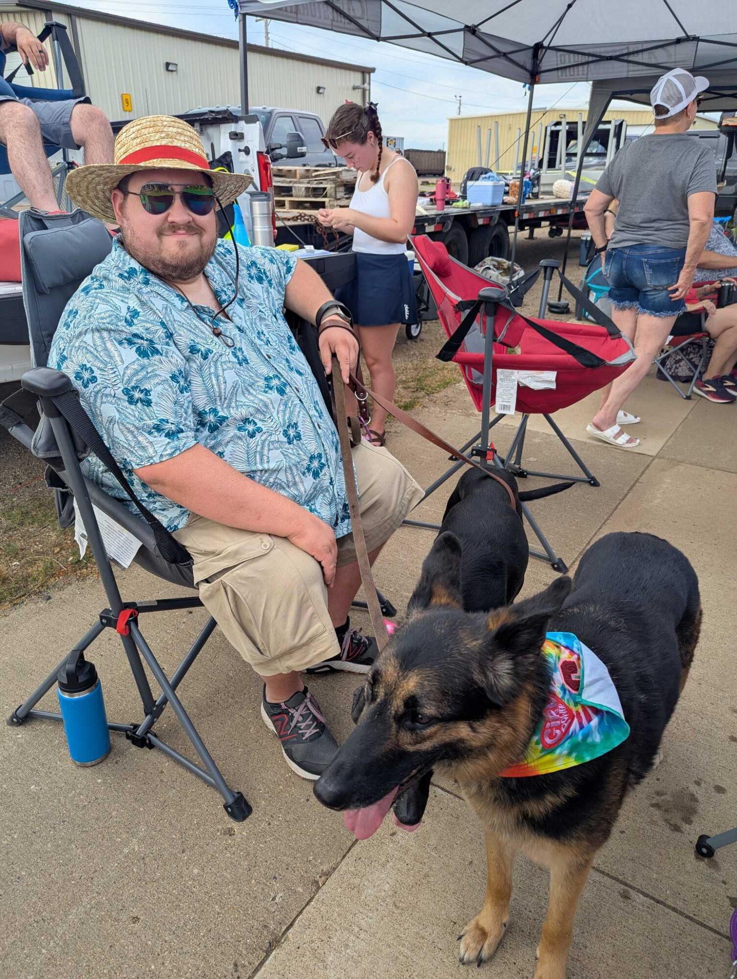 A man wearing a Hawaiian shirt and straw hat sits in a folding chair in the street, it appears he is watching a parade. He is holding two dogs on leashes, one is wearing a tie-dye bandana around their neck.