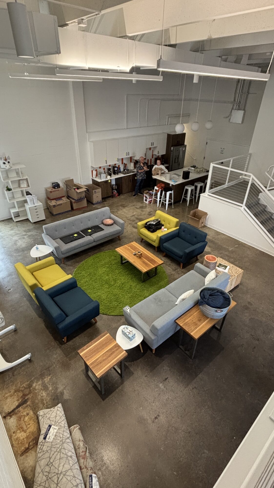 A loft-style lounge area is shown from above, featuring colorful mid-century modern furniture arranged around a green circular rug. The space includes a kitchen with white cabinets and barstools, and two people are seen chatting near the counter. Cardboard boxes are visible, suggesting the space is being unpacked or rearranged.