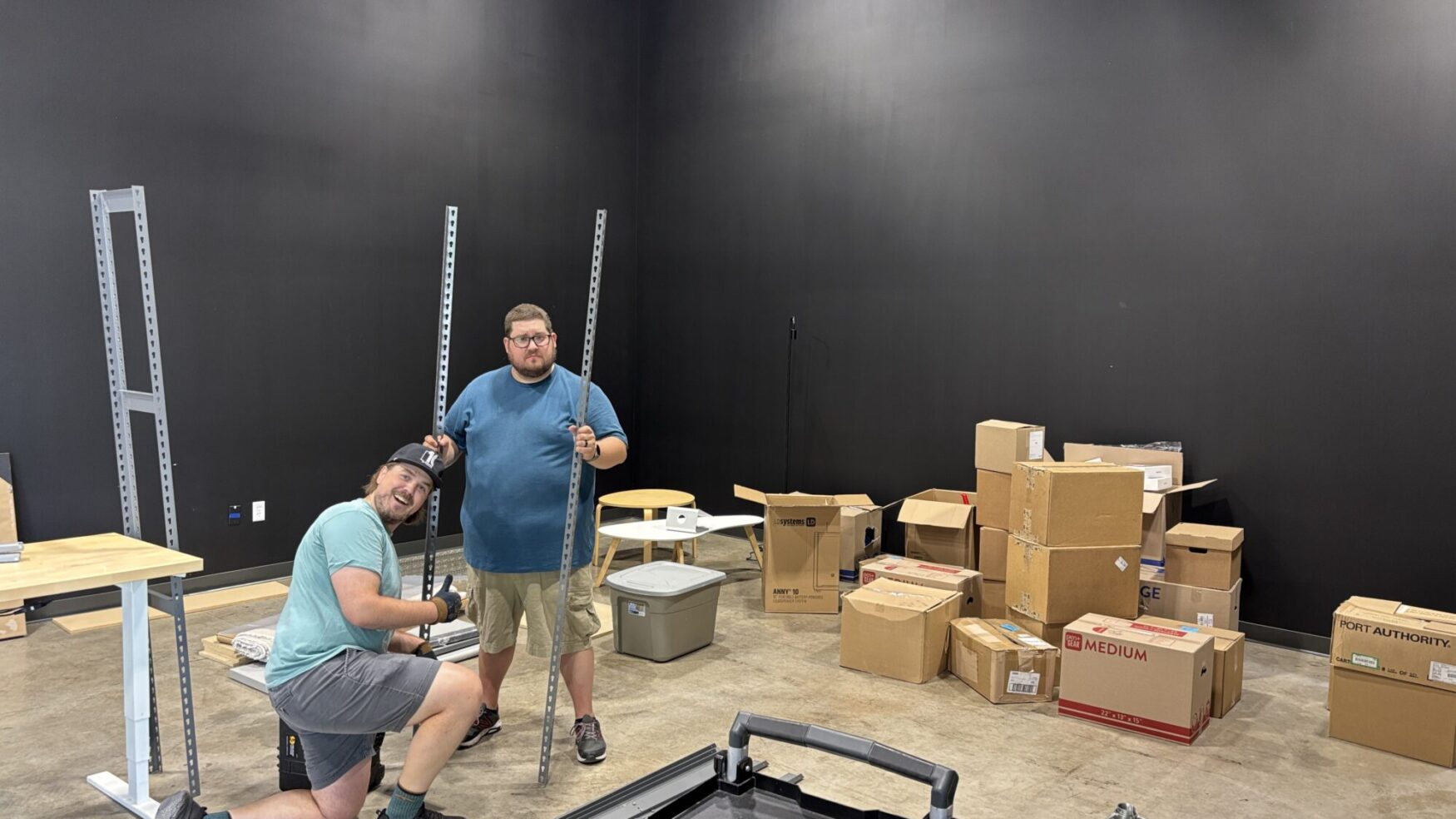 Two men are assembling metal shelving in a nearly empty room with a black wall. One kneels with a tool, smiling at the camera, while the other stands holding a metal frame. Several cardboard boxes and various packing materials are scattered around the room, indicating a move-in or setup process.