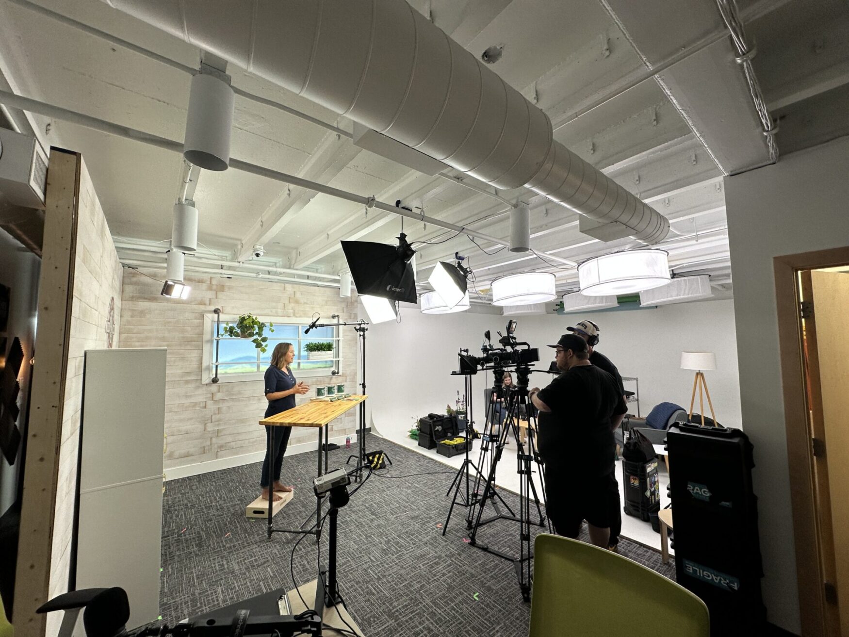 A behind-the-scenes look at a video shoot in a studio space. A woman stands behind a wooden table, speaking on camera under professional lighting. Crew members operate cameras and equipment. The set features light wood paneling, fake windows with a scenic backdrop, and houseplants, creating a clean, inviting scene.