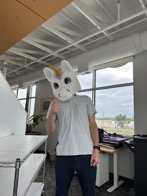 A person wearing a large, whimsical unicorn head costume gives a thumbs-up in a brightly lit office with high ceilings and large windows. The individual wears casual clothes—jeans and a gray T-shirt. Office furniture and equipment are visible in the background, giving the space a creative, informal feel.