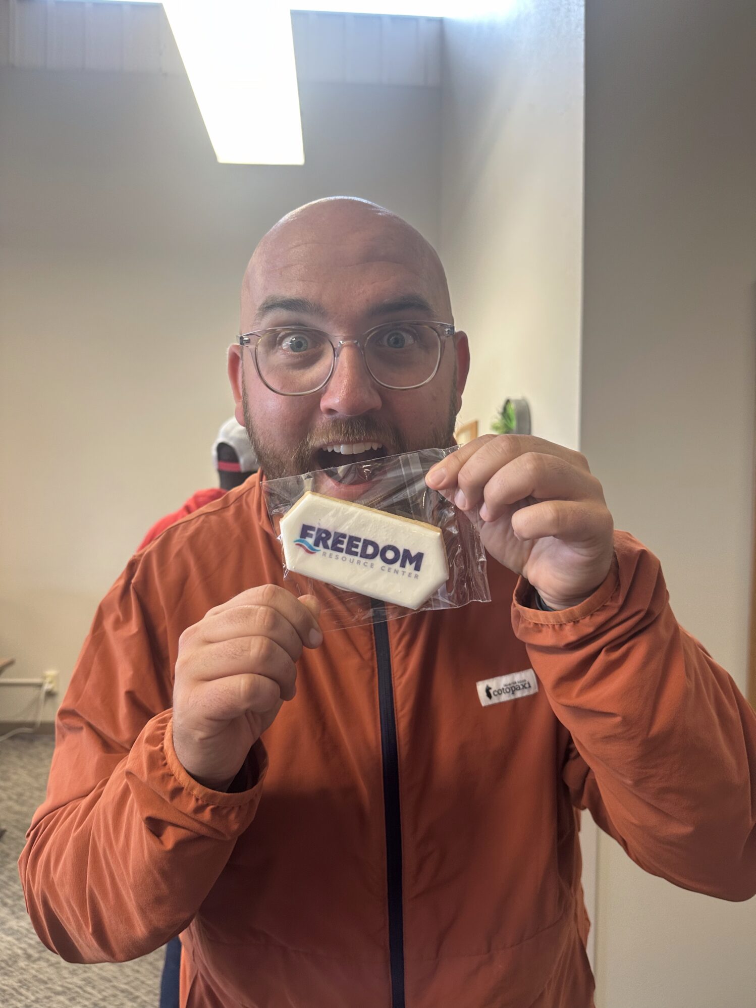 A man in an orange jacket holds a bag with a cookie inside. The cookie is white and reads "Freedom Resource Center". He looks excited, with his eyebrows raised and his mouth open.