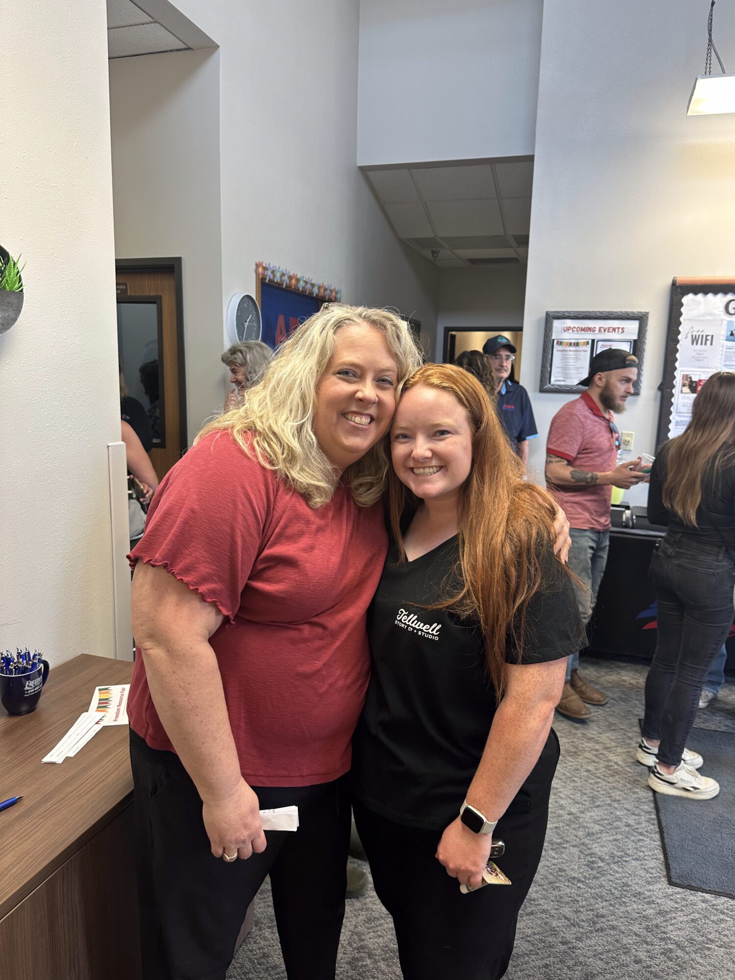 Two women stand close together for a photo together. They are in an office setting with several other people in the background, suggesting an office gathering is happening.